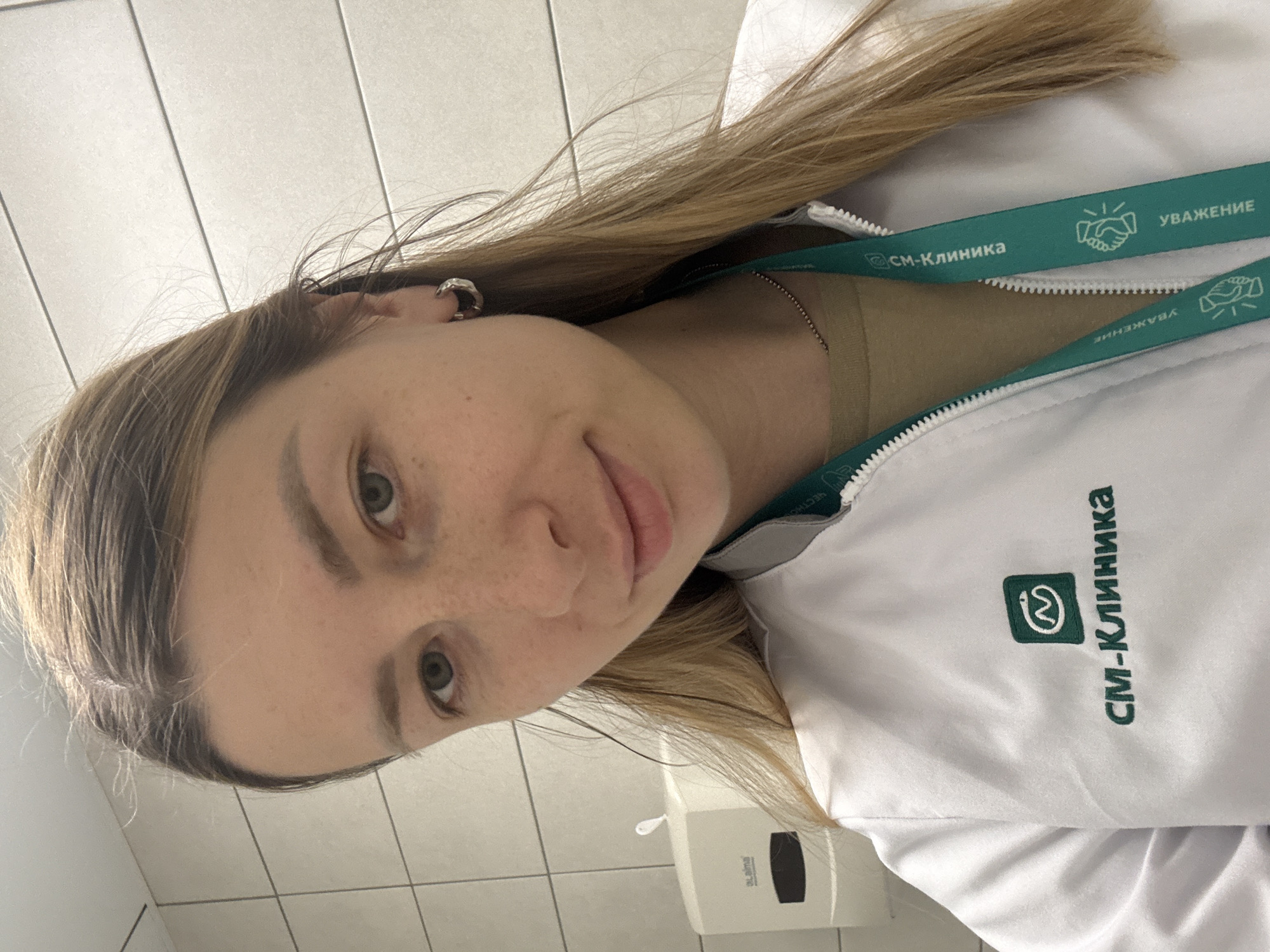 The image shows a close-up, waist-up selfie of a young woman wearing a white lab coat with a teal lanyard. The lab coat has a green logo with text that reads "СМ-Клиника" (SM-Klinika). The lanyard also has the same logo and text, along with other graphics. The woman has long, blonde hair and is wearing a silver hoop earring. In the background, there is a white paper towel dispenser mounted on a tiled wall. The setting appears to be an indoor medical or clinical environment. Without further information, the specific location in Moscow, Russia cannot be determined.