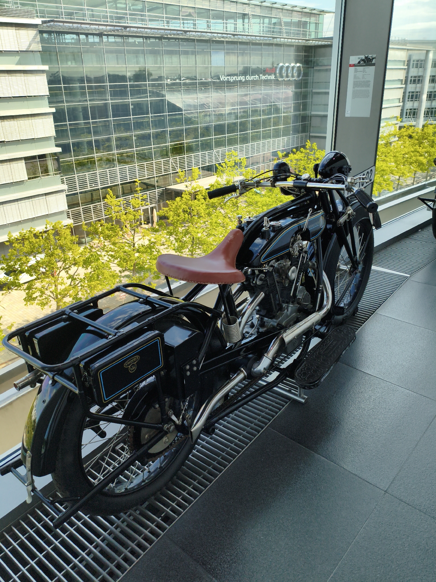 The image depicts a vintage black motorcycle displayed indoors, with a modern glass building visible through a large window in the background. The motorcycle is positioned on a metal grate flooring, with its rear wheel closest to the viewer. It features a brown leather seat, a luggage rack at the back, and chrome exhaust pipes. A license plate with the number "19022" is attached to the rear fender.
The building in the background is the Audi Forum in Ingolstadt, Germany. Its facade is characterized by large glass panels and horizontal silver louvers. The "Vorsprung durch Technik" slogan and the Audi logo are clearly visible on the building's exterior. Lush green trees with bright yellow foliage are seen through the window, suggesting it is springtime or early summer. The lighting indicates an overcast or bright day. There are no people visible in the image.