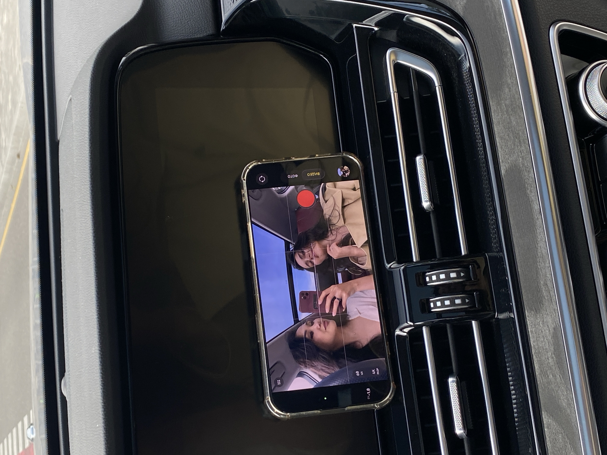 An iPhone, displaying a video call between two women, rests on the dashboard of a car. The dashboard is black, featuring a rectangular tray and an adjacent air vent with chrome accents. The car is stationary, as suggested by the roadside visible through the windshield. The location is unclear, as there is no visible information indicating Sirius, Russia.