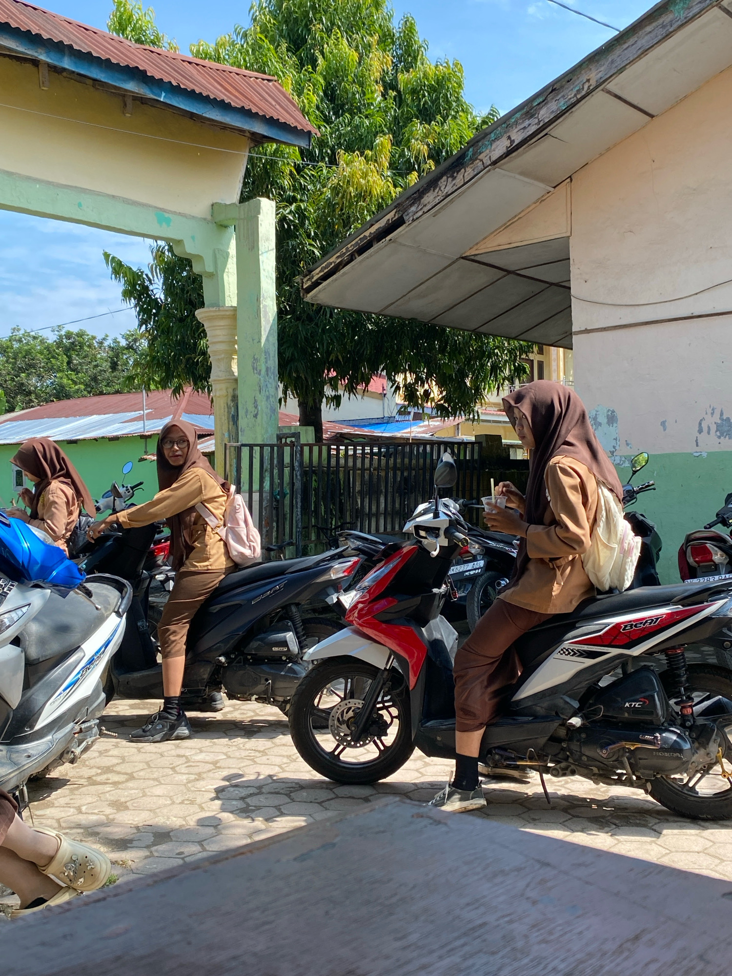 In Karang Gading, Indonesia, three young individuals, two clearly identifiable as female students wearing matching brown uniforms and headscarves, are seen around parked motorbikes. One student, positioned on a black motorbike, is holding a cup and a straw. Another student, on a different black motorbike, is also seated, facing away from the camera. A third student, partially visible in the foreground, is seated on a scooter with a blue covering. The scene unfolds outdoors with a building featuring a green and yellow facade and a rustic metal roof in the background, surrounded by lush green trees under a bright, clear blue sky. The ground is paved with interlocking stones, and a shadow from a table or bench falls across it.