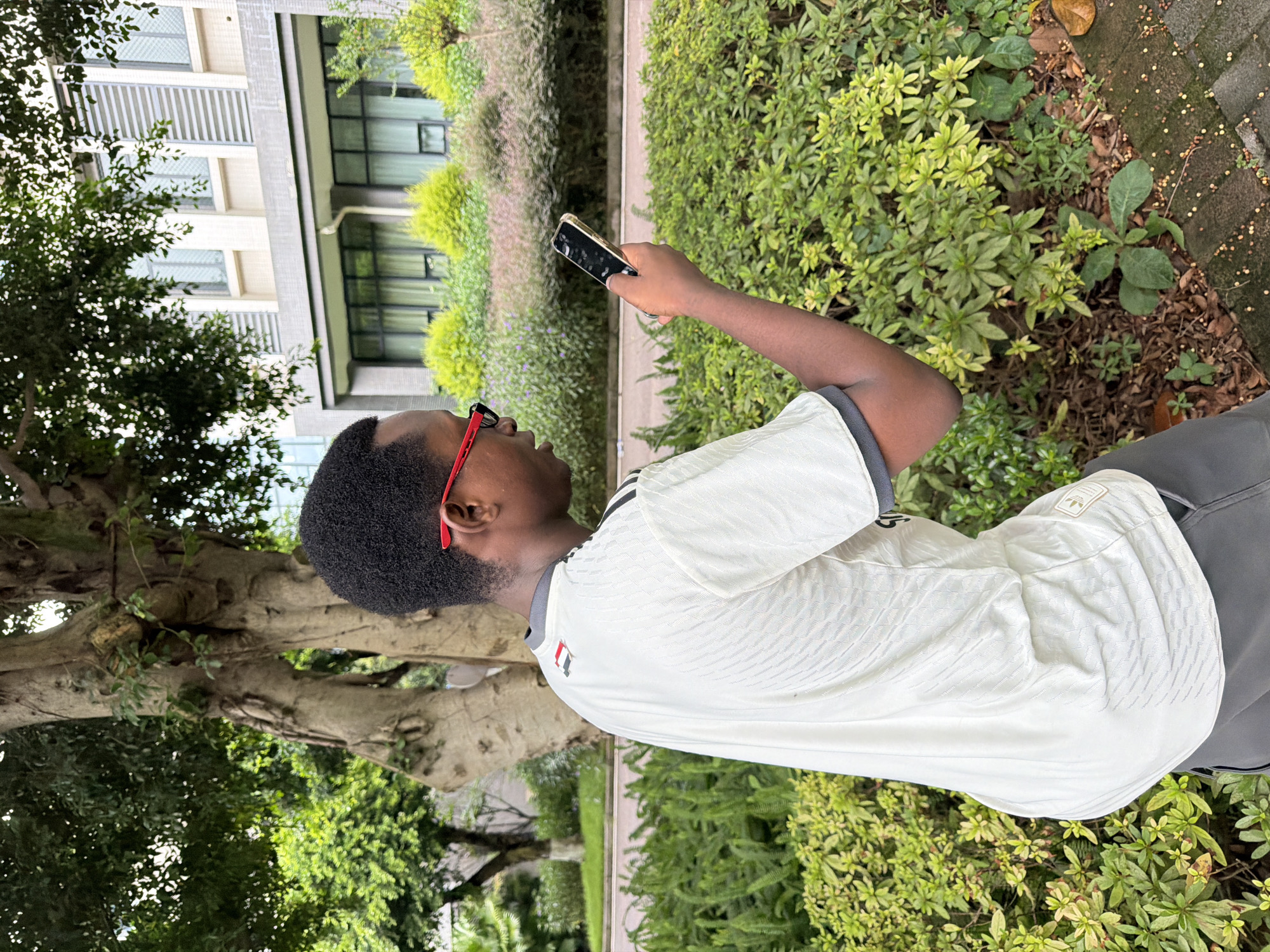 A young Black male wearing a white t-shirt with black accents and gray shorts stands outdoors, holding a smartphone in his right hand. He is looking down at the phone. The background features a large tree on the left, a building with multiple windows and horizontal blinds in the middle, and lush green bushes and plants. The scene appears to be in a park or a well-maintained outdoor area. The context of the image being taken in Jiangjin, China, suggests it is a location within that city.