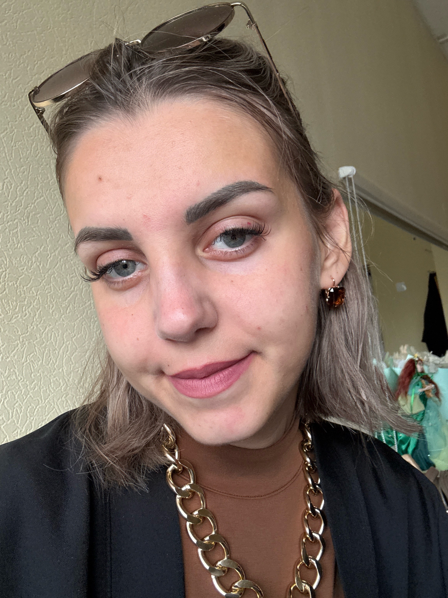 A young woman with light brown hair, pulled back and resting on her shoulders, is captured in a close-up, head-and-shoulders shot. She is wearing a brown turtleneck and a black blazer, accented by a thick gold chain necklace. Her makeup includes mascara, eyeliner, and pink lipstick. She also sports large, dark brown earrings and sunglasses perched atop her head. Her gaze is directed slightly to the left, and she appears to be indoors, with a textured beige wall behind her. The background also reveals a hint of a mirror and some decorative items. The lighting is soft and natural. The image appears to be a selfie taken in Yuzhno-Sakhalinsk, Russia.