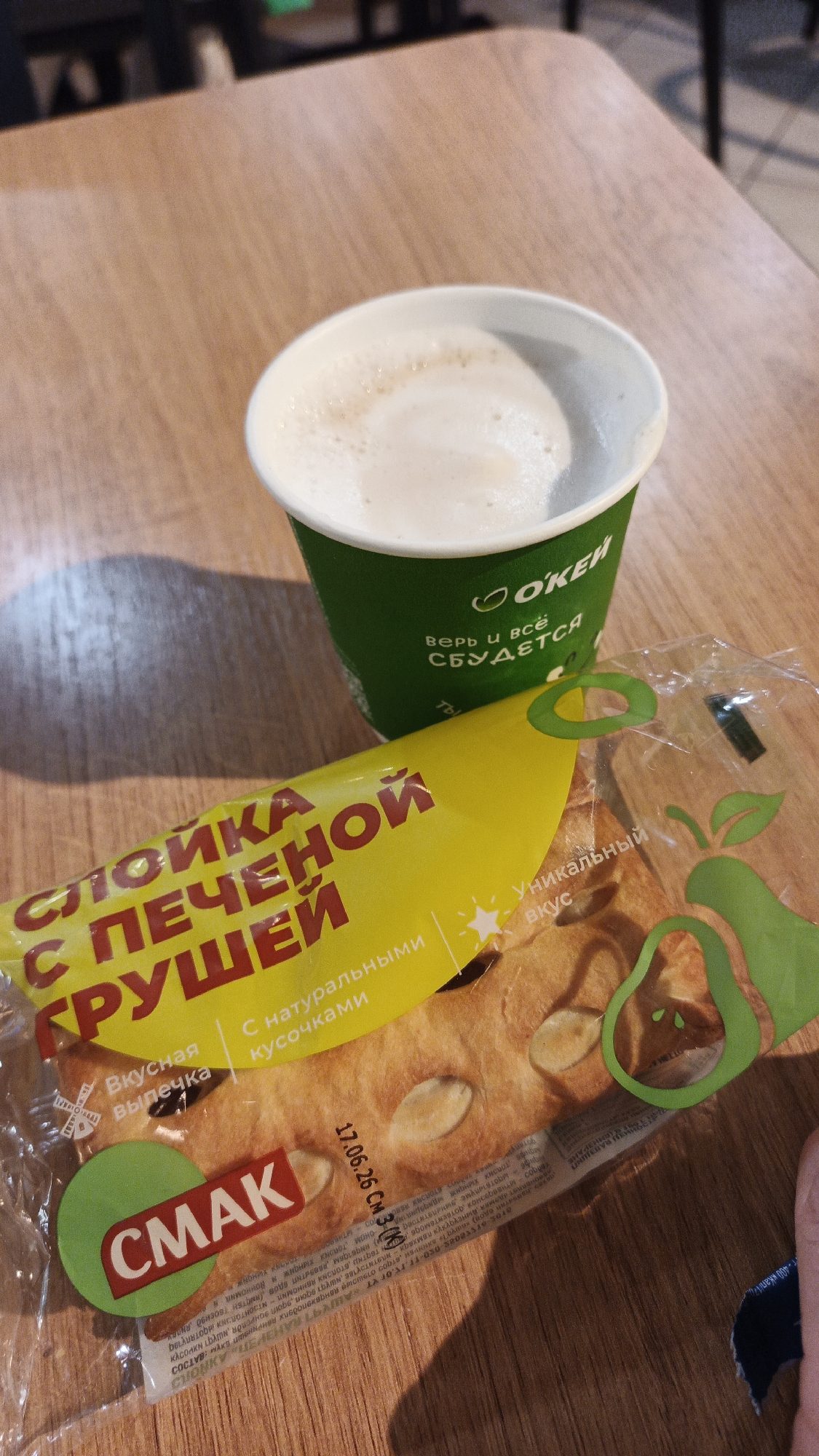 The image depicts a close-up, eye-level view of a wooden table with a green paper cup filled with a frothy beverage, likely coffee, and a packaged pastry. The cup, bearing the "O'KEY" logo and Russian text that translates to "Believe and everything will come true," is positioned in the upper right portion of the frame. A yellow wrapper, also with Russian text indicating "Puff pastry with pears," is placed diagonally across the lower half of the image, partially obscuring a pastry inside the clear plastic packaging. The pastry appears to be a flatbread or cookie topped with what look like slivers of almonds. A red circular logo with the word "SMAK" is visible on the lower left of the wrapper. Faint text and a date, "17.06.26," are visible on the packaging. The lighting suggests an indoor setting, possibly a cafe or supermarket, with soft shadows cast on the table. No specific geographical markers are present to indicate the city of Yekaterinburg, Russia.