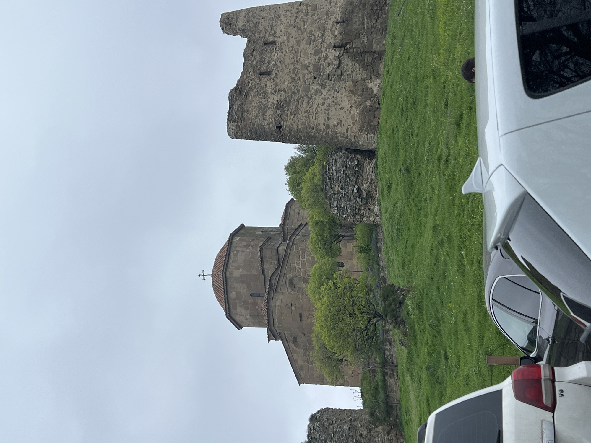 The image shows the Jvari Monastery, located near Mukhatgverdi, Georgia. The monastery, a stone building with a dome topped by a cross, stands on a hill with green grass and some trees. In the background, there is a stone tower ruin. In the foreground is a portion of the side and rear of two white vehicles, partially obscuring the scene. The sky is overcast.