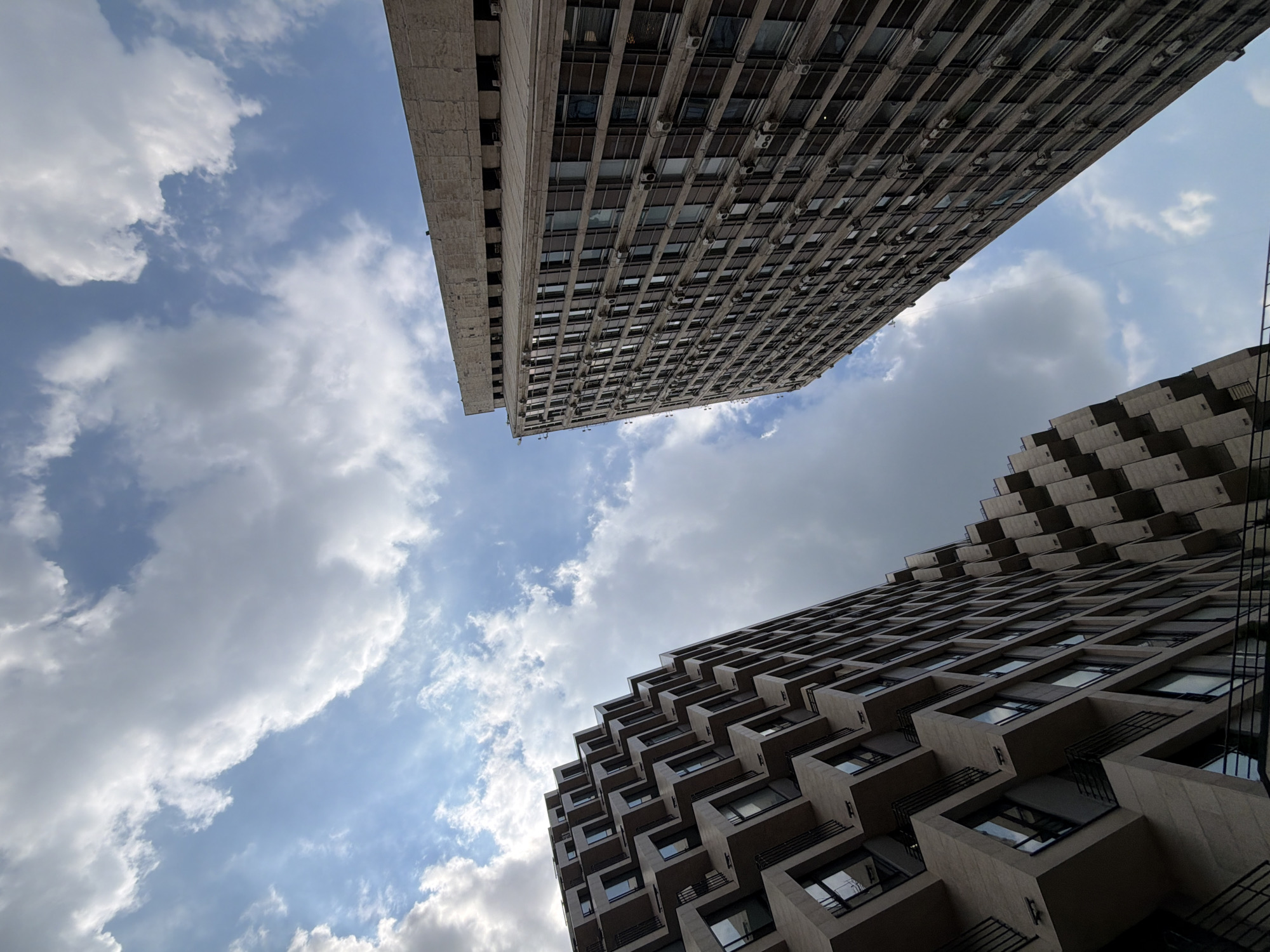 Two modern high-rise buildings, one with a distinctive stepped facade and the other a tall, rectangular structure, are captured from a low-angle perspective against a partly cloudy sky. The building on the left features a repeating pattern of protruding, cube-like balconies with dark railings. The building on the right is a sheer, multi-story structure with numerous windows and visible air conditioning units. The sky is a vibrant blue with scattered white and grey clouds. The scene appears to be an urban environment, likely in Moscow, Russia, given the architectural style and commonality of such structures in major Russian cities. No people are visible in the image.