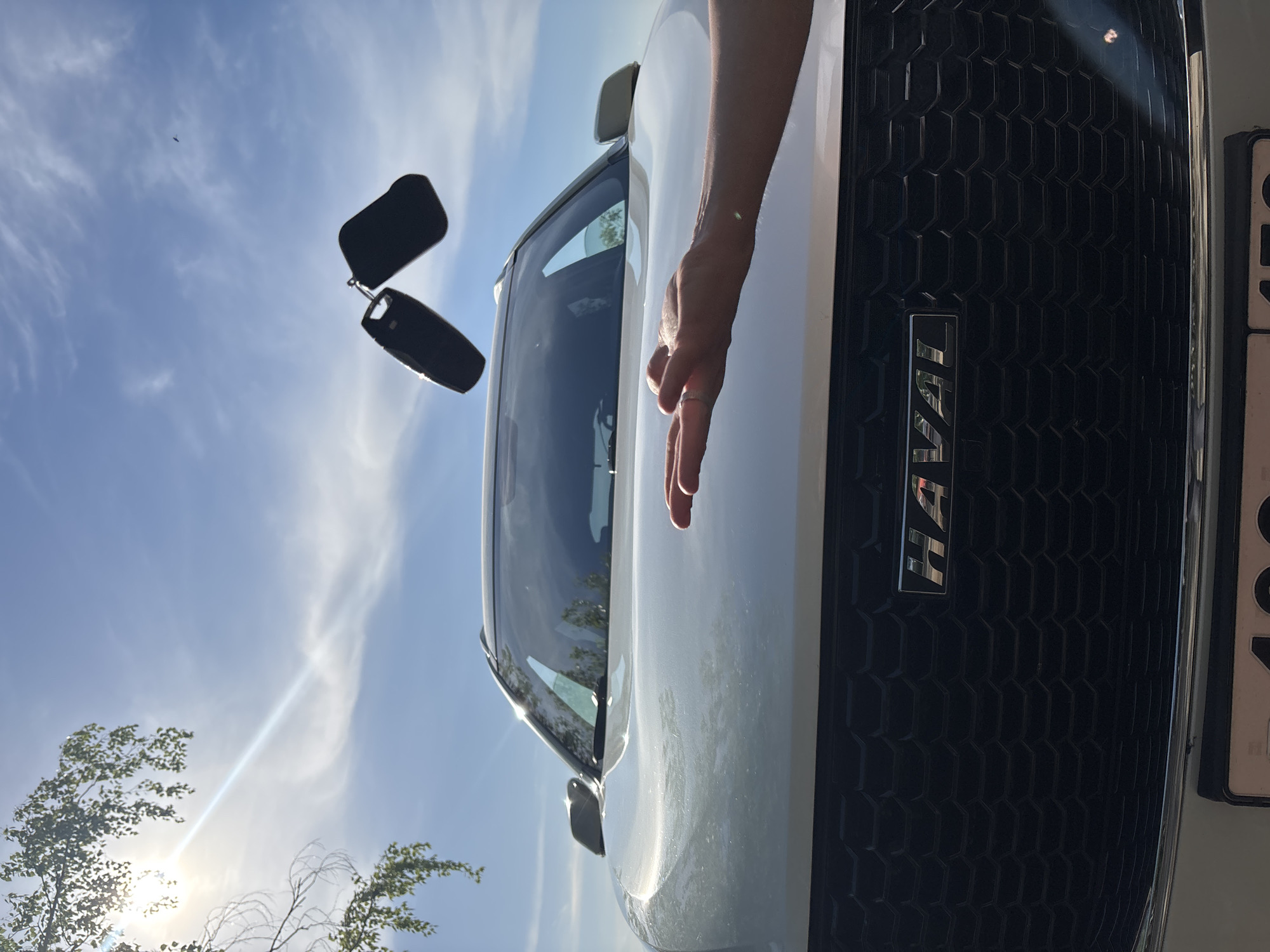 The image shows a close-up of the front of a white Haval car, with a person's hand resting on the hood. The car keys are suspended in mid-air against a blue sky with some clouds. The car's grille and Haval logo are visible. The license plate is partially obscured. A small tree is visible in the lower-left corner of the frame. Based on the information provided, the location is Chaikovsky, Russia.