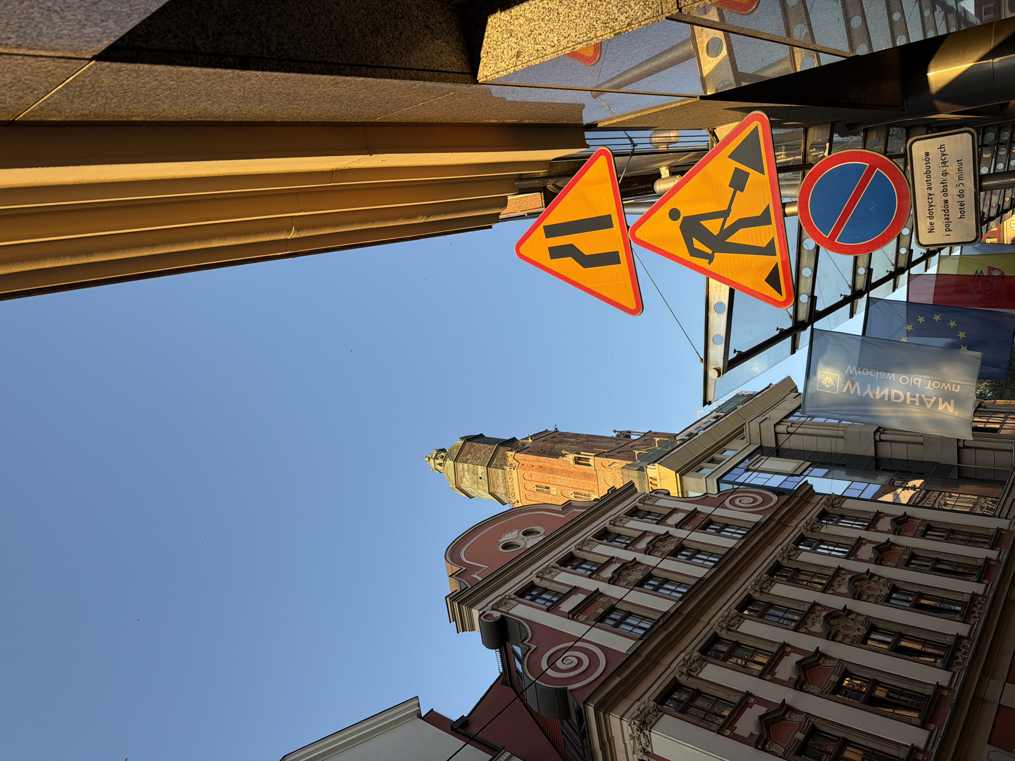 The image captures a street scene in Wrocław, Poland, on a clear, sunny day with a bright blue sky. In the foreground, several road signs are visible. The topmost sign is a yellow triangle with a red border, depicting a black arrow indicating a narrowing of the road from the right. Below it is another yellow triangle with a red border, featuring a black silhouette of a person digging with a shovel, signifying road work. Beneath these is a circular blue sign with a red border and a red diagonal line, which is a prohibition sign, likely meaning "no stopping" or "no parking." A small rectangular white sign with black text in Polish is attached below the prohibition sign.
To the left of the signs, an ornate, multi-story building with a reddish-pink facade and white architectural details stands prominently. It features numerous windows and intricate ornamentation. Further in the background, a tall, brick clock tower with a greenish-bronze dome rises into the sky, characteristic of historical European architecture. The building to the right of the signs appears more modern, with a dark, reflective facade and a golden, ribbed architectural element extending upwards. Flags, including what appears to be the European Union flag, are also visible hanging behind the signs. The overall impression is a blend of historical and modern urban elements under a clear sky.