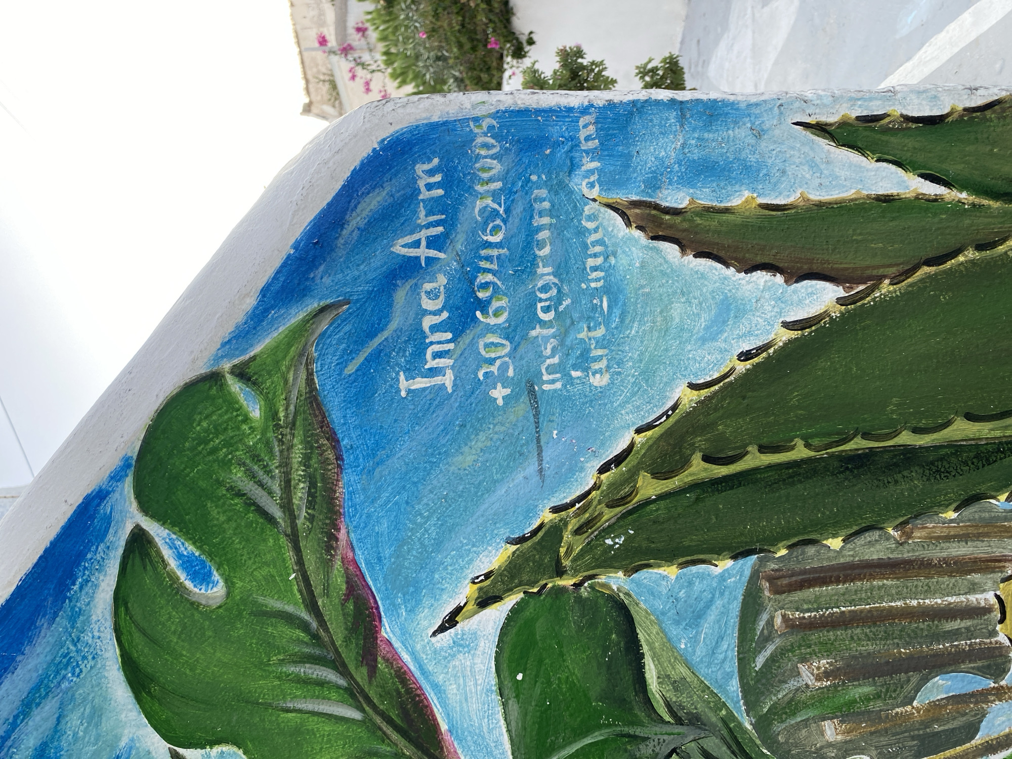 The image shows a close-up view of a painted mural on a curved surface, likely a wall or a sculpture, in Thira Municipal Unit, Greece. The mural features stylized green leaves, possibly representing aloe vera or similar succulents, against a bright blue sky with white brushstrokes. White text is painted onto the blue background, indicating contact information. The text reads "Inna Arm", "+30 694621005", and "instagram: art-innoarm". In the background, out of focus, is a white building and some pink flowers, typical of a Mediterranean setting. The lighting suggests it is daytime, with the sun casting a bright light on the scene.