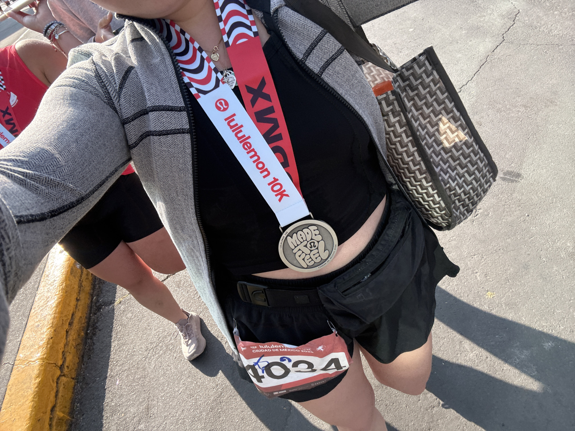 A woman wearing a black crop top, black shorts, a gray patterned zip-up jacket, and a black fanny pack is seen holding a medal from a Lululemon 10K race in Mexico City. The medal, emblazoned with "Made to Feel," hangs from a ribbon. The race bib number "4024" is attached to her shorts. She also has a large tote bag over her shoulder. Another person wearing a race tank top and shorts is partially visible to the left. The setting appears to be on a city street in Mexico City, Mexico, as indicated by the medal and bib information. The ground is paved.