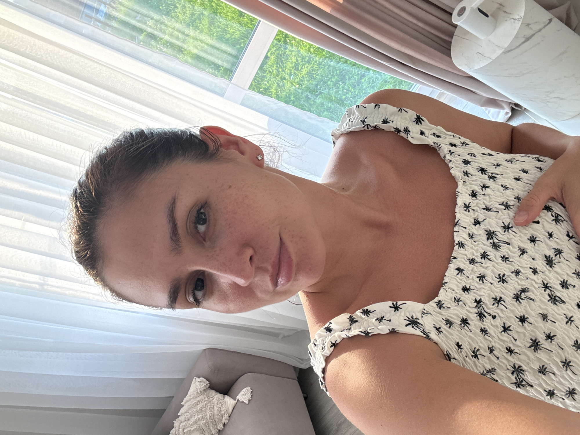 This is a first-person, vertical, medium shot of a woman in her home. She is looking directly at the camera with a neutral expression. Her hair is pulled back, and she has freckles on her face. She is wearing a white top with a black palm tree pattern. The background shows a window with white blinds and lush green grass visible outside, suggesting it is daytime and likely warm weather. To the right of the woman, a modern-looking white lamp or diffuser sits on a surface with a marble-like pattern. A grey armchair with a textured white pillow is visible in the lower left portion of the image. The overall scene suggests a relaxed, indoor setting. The time of day appears to be daylight, with bright sunlight streaming through the window. The location context provided is Нижнеяркеево, Russia. There is no visible text in the image.