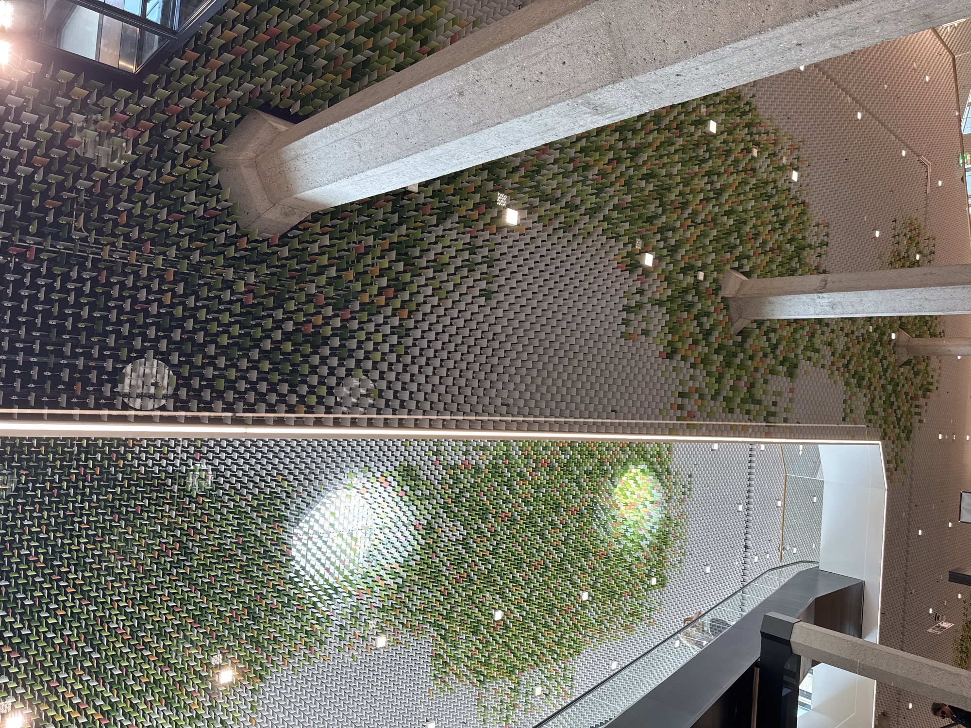 The image depicts the interior of a modern building, likely a commercial or public space, with a striking ceiling design. The ceiling is covered in a multitude of small, square-shaped tiles arranged in a grid pattern. Many of these tiles are colored in shades of green, orange, and red, giving the impression of foliage or a canopy. In some areas, the tiles are denser and more vibrant, suggesting lush vegetation, while other sections are more sparsely populated or predominantly grey, creating a textured background.
Illuminating the space are recessed square lights embedded within the ceiling grid. Reflections of these lights and the surrounding environment are visible on some of the metallic surfaces, particularly on the left side of the image where a reflective panel or ceiling section is present.
Prominently featured are two large, cylindrical concrete pillars supporting the structure. The texture of these pillars is rough and unfinished, contrasting with the more refined ceiling elements.
To the left, a portion of a multi-level structure is visible, including a sleek grey railing and what appears to be the edge of an escalator or staircase. A few individuals are present in the scene. On the far left, a person's shoulder and upper arm, wearing a dark garment, are visible. Further up on the railing, a person is seen ascending on an escalator. In the lower right corner, a person wearing sunglasses and a dark top is partially visible.
The overall environment suggests a contemporary architectural design that incorporates natural elements and light. The mention of Morbio Inferiore, Switzerland, implies this location is within a building situated in that Swiss municipality.