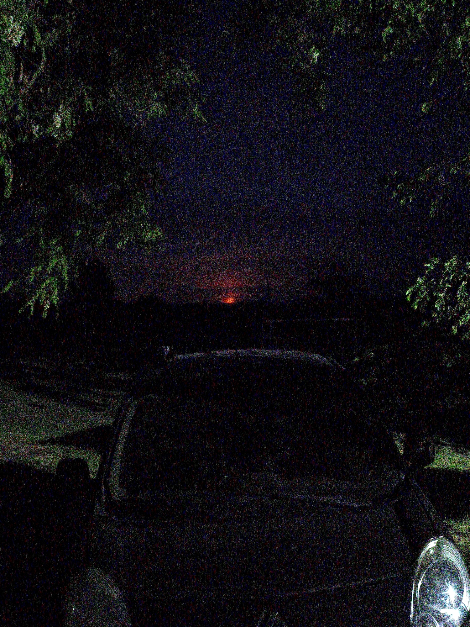 The image shows a car parked outside at night. The car's hood and windshield are visible in the foreground. Behind the car, the sky is a deep blue with a hint of red or orange low on the horizon, possibly indicating a sunset or sunrise. Trees surround the car on both sides. The image has a grainy texture.