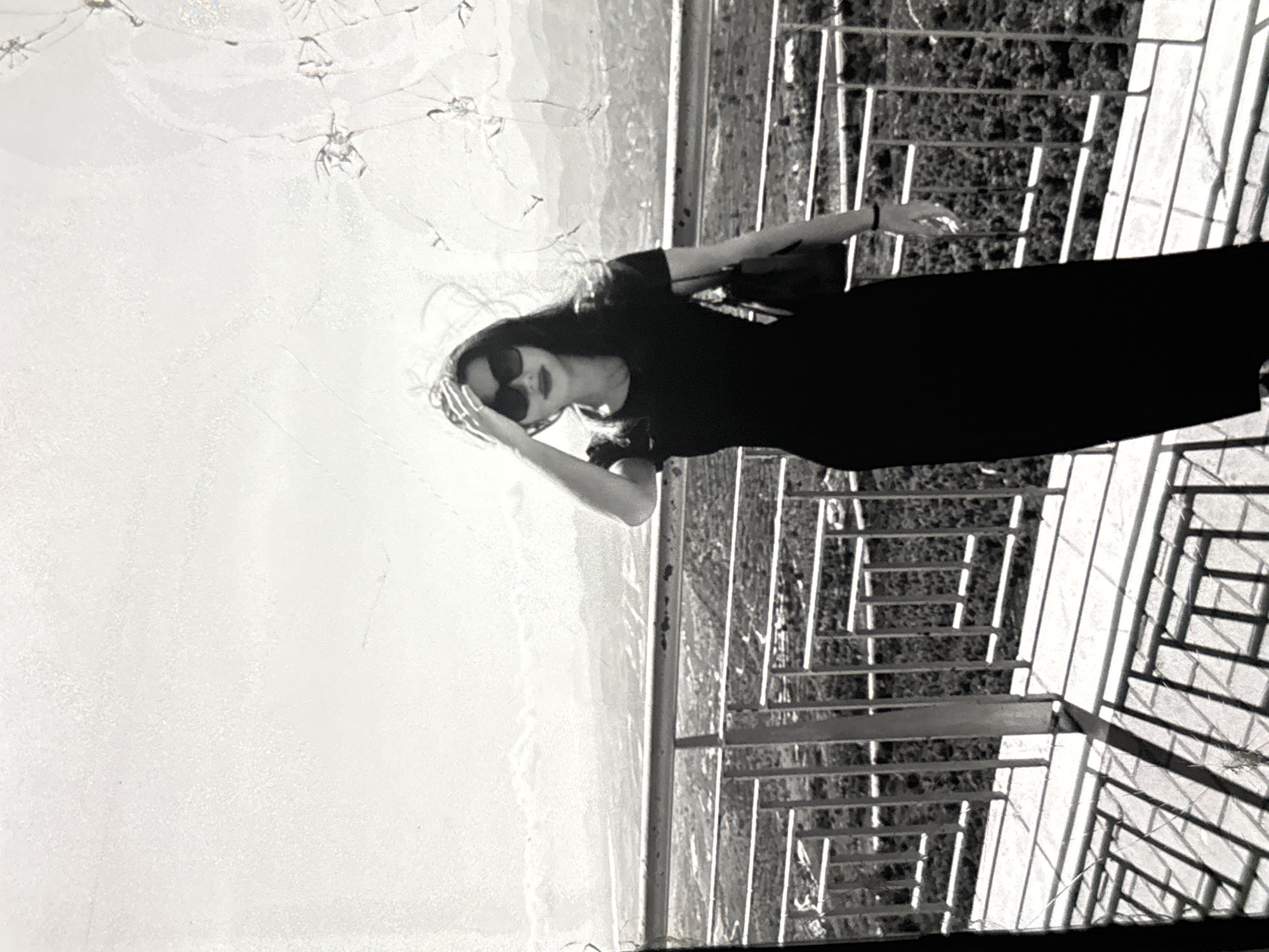 A black and white, grainy photograph shows a woman standing on a balcony overlooking a landscape. She is wearing a dark, floor-length dress and sunglasses, with one hand touching her hair. Behind her, a metal railing separates the balcony from the view of trees and a distant city. The sky is bright and overcast. The photograph has a distressed appearance with visible cracks and scratches overlaying the image. The location is identified as the city of Orihuela, Spain.