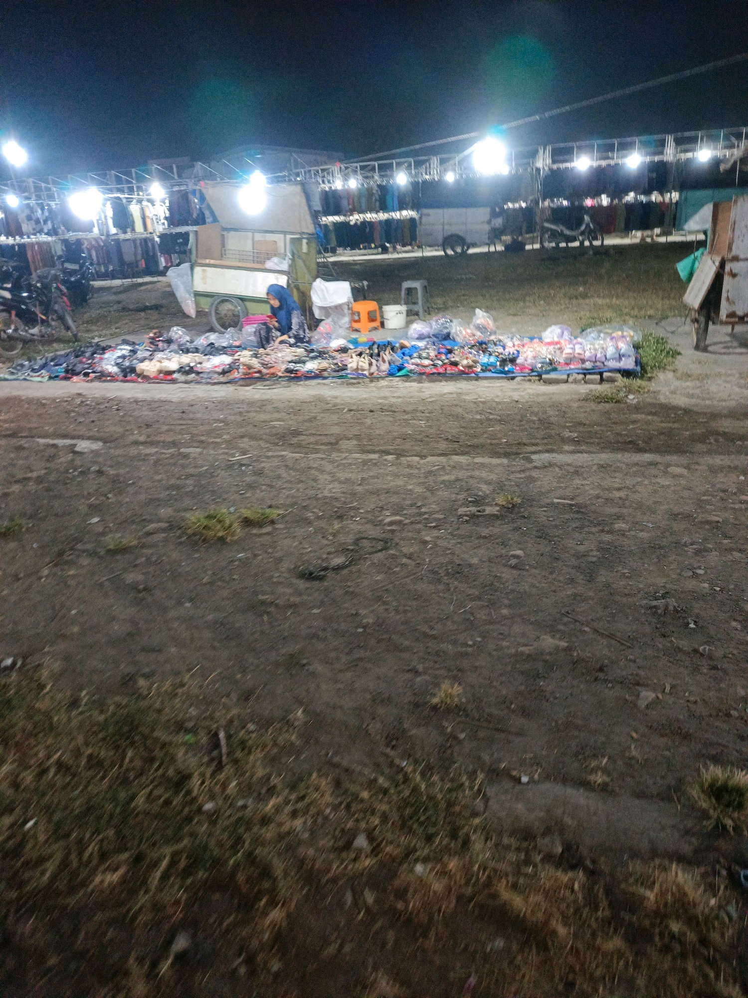 The image depicts an outdoor market at night, likely in Sedati Agung, Indonesia, illuminated by bright overhead lights. In the foreground, a large spread of various footwear, including sandals and flip-flops, is displayed on the ground, arranged on what appears to be tarps and cloths. Some of the footwear is in plastic bags. A person wearing a blue hijab is seated amongst the footwear, appearing to be sorting or arranging it. Behind the footwear display, a light-colored cart with a canvas cover is visible, with a person in a green shirt standing near it. Further back, rows of clothing racks are set up under a canopy structure, with garments hanging on display. Motorcycles are parked to the left of the clothing racks. The ground is a mix of dirt and patchy grass. The overall scene suggests a busy night market environment.