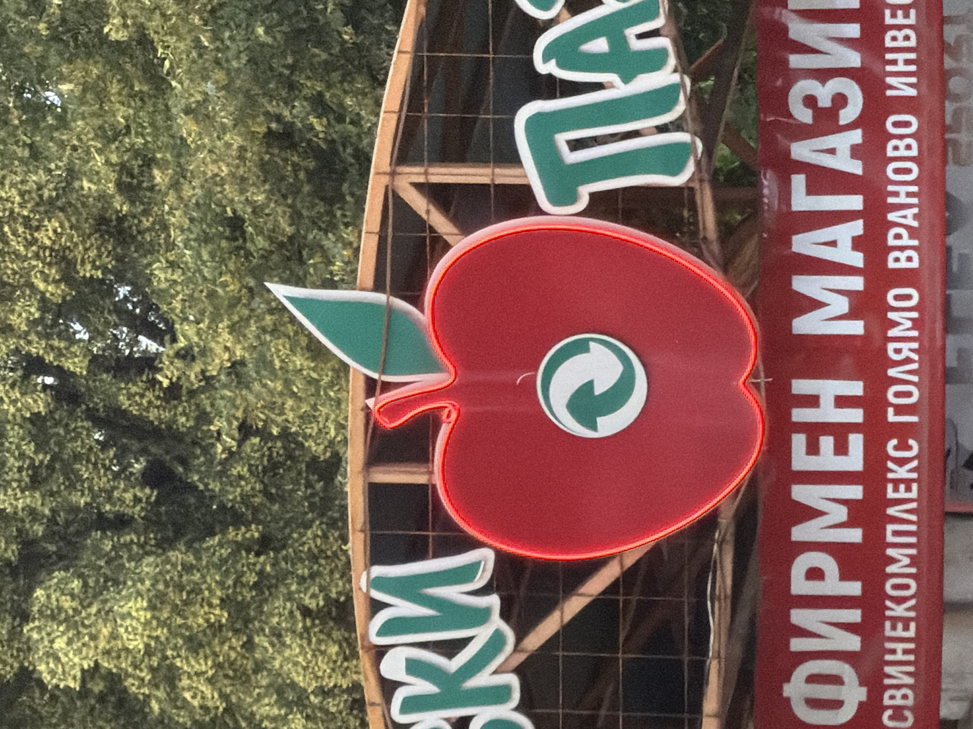 The image shows a sign with an apple logo and text in Bulgarian. The sign is red and green with white lettering. The apple logo is red with a green leaf. Inside the apple, there is a white circle with two green arrows forming a recycling symbol. The text on the sign reads "ФИРМЕН МАГАЗИН" (Firm Store) on the top line and "СВИНЕКОМПЛЕКС ГОЛЯМО ВРАНОВО ИНВЕСТ" (Pig Complex Golyamo Vranovo Invest) on the bottom line. The sign is mounted on a metal structure and is set against a background of green trees. This sign is located in the city of Ruse, Bulgaria, specifically on "Tsar Ferdinand" street, near the "Ruse-West" highway entrance, in front of the "Golyamo Vranovo" pig complex.