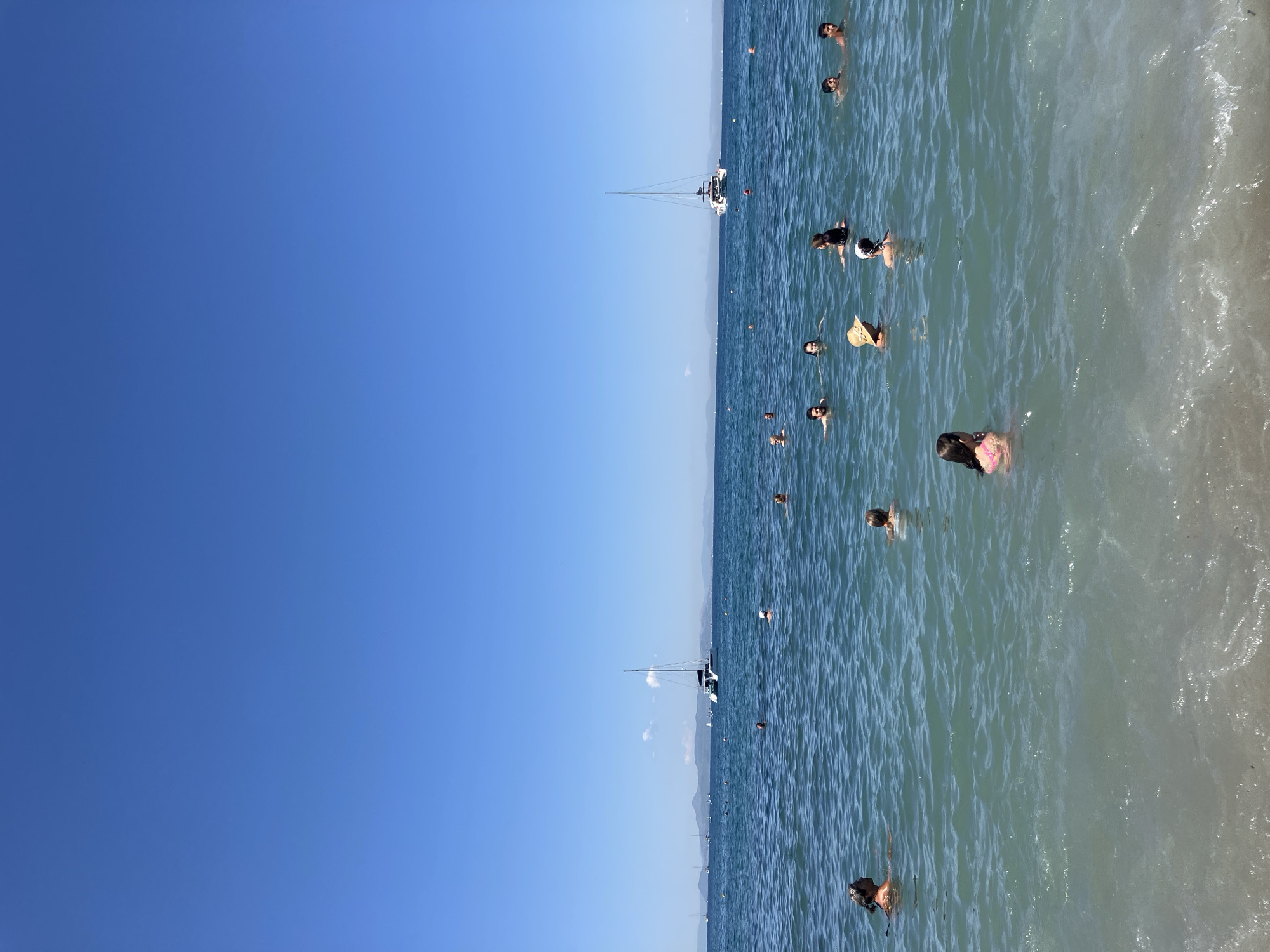 This is a wide-angle, eye-level shot of people swimming in the sea in Paleo Faliro, Greece, on a clear, sunny day. The water is a mix of turquoise and deep blue, with some light chop and a sandy seabed visible closer to the shore. In the background, a large white catamaran with a single mast is sailing, and several other smaller boats and yachts are dotted across the horizon. Distant mountains form a hazy line against the bright blue sky. A few buoys are visible in the water. Several people are scattered throughout the frame, mostly immersed up to their shoulders or necks. A woman in a pink bikini is in the foreground, facing away from the camera. Another person wearing a straw hat is to her right. The overall atmosphere is one of leisure and recreation.