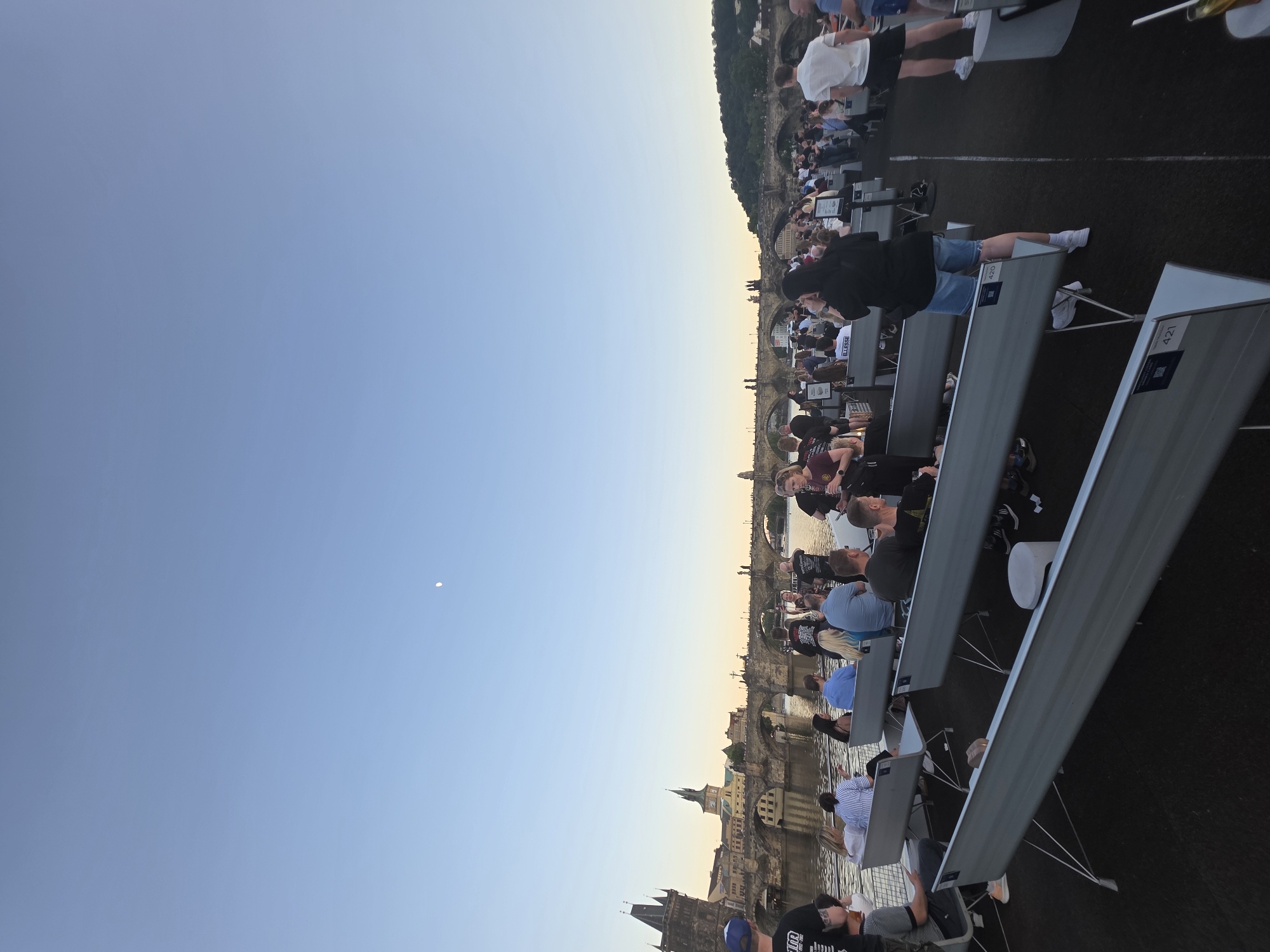 The image shows a daytime outdoor scene in Prague, Czech Republic, with a view of the Charles Bridge and the Vltava River. A crowd of people are seated in rows of chairs with attached desks, possibly attending an event. The sky is blue with a hint of orange near the horizon, and a small moon is visible. The architecture of the city, including towers and buildings, is visible in the background along the riverbank.