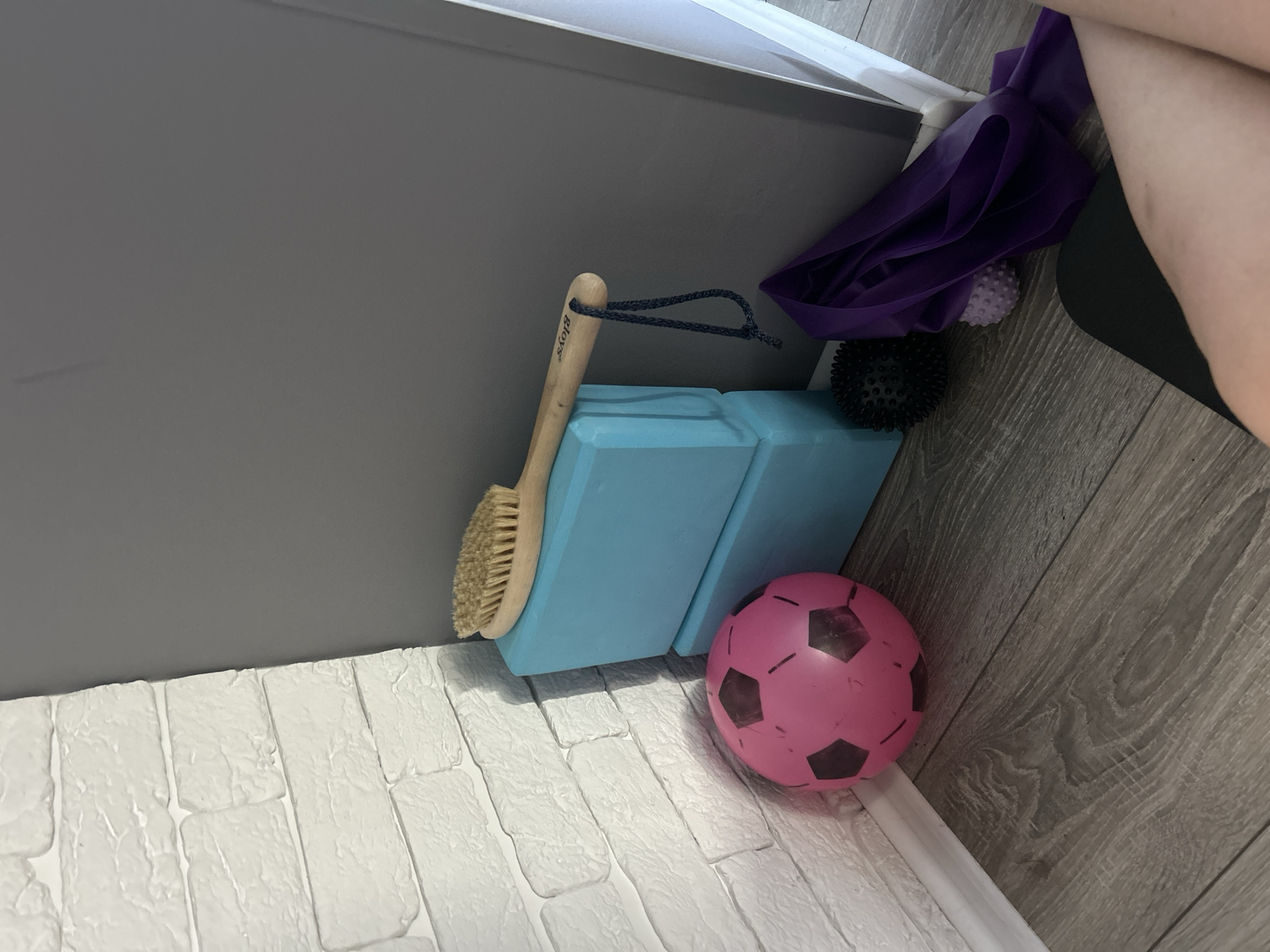 An indoor scene in Orenburg, Russia features a collection of fitness equipment in the corner of a room. The items are placed on a grey wooden floor next to a white brick-patterned wall and a dark grey wall. On the floor, a pink soccer ball with black pentagons and a black spiky massage ball are visible. A purple resistance band is draped nearby, partially obscuring a small, light purple textured ball. Stacked on top of each other are two bright blue yoga blocks. Resting on the upper yoga block is a wooden-handled brush with natural bristles, a dark blue string loop attached to its handle. The lighting suggests an interior environment, possibly a home gym or a fitness studio.
