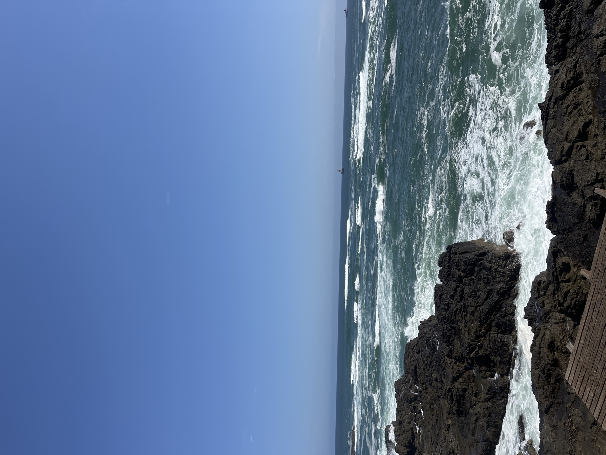 The image shows a coastal scene in Arica, Chile. The sky is clear blue, occupying half of the image. The other half depicts the ocean and a rocky shoreline. The ocean waves are white and foamy. Dark, volcanic rocks form the coastline. A partial view of a wooden structure is seen on the right edge of the frame. Distant ships are visible on the horizon.