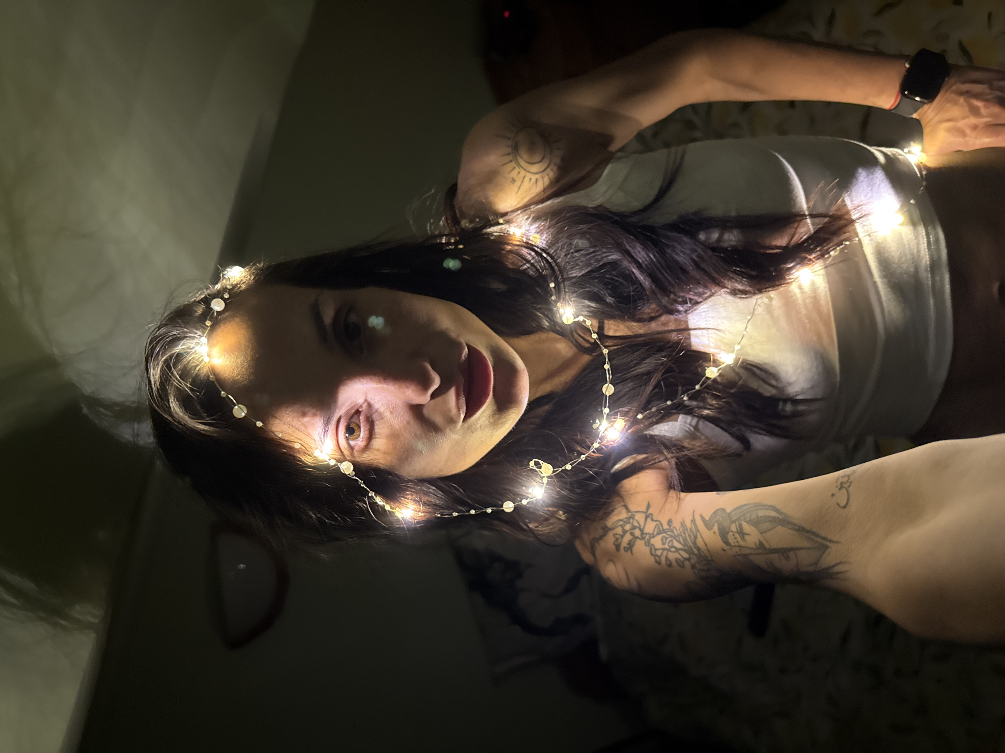 A woman with long, dark hair is posing indoors. She is wearing a white tank top and has a string of small, white lights wrapped around her head and torso. She has tattoos on both arms. One tattoo appears to be a sun and moon design, and the other is a more detailed, dark ink design. She is wearing a black smartwatch on her wrist. The background is dark and partially obscured, but a wall and the edge of what might be a bed are visible. The photograph appears to be taken in low light. There is no indication of this image being in Pleven, Bulgaria.