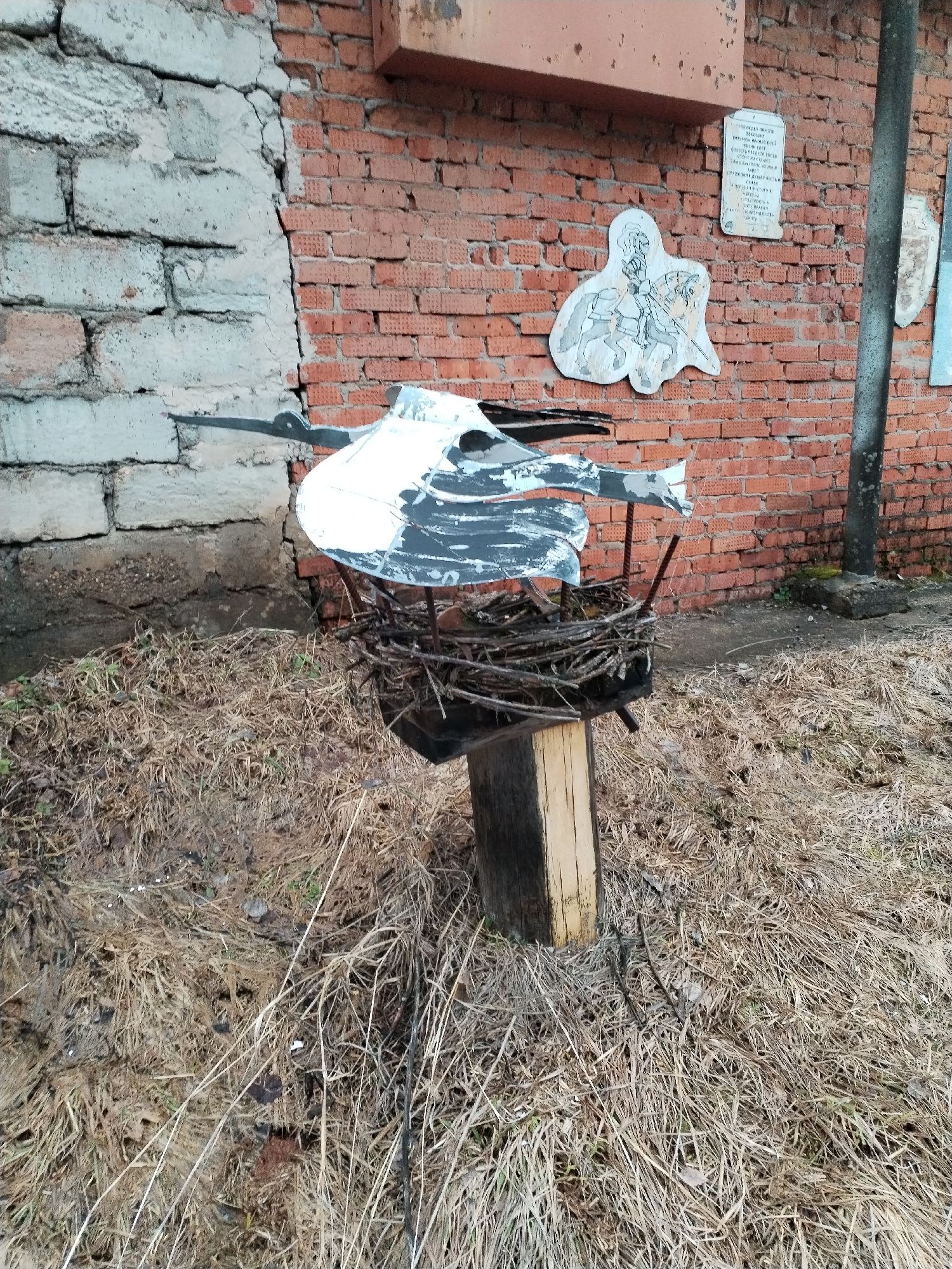 The image shows a handcrafted stork decoration perched atop a wooden post. The stork is made of painted metal and sits in a nest of twigs. The post is planted in an area covered with dry grass and weeds. Behind the decoration, there is a brick wall partially covered with crumbling concrete. On the brick wall are several plaques and painted decorations. The location appears to be an urban environment, based on the presence of the brick wall and other structures. The city and region are unknown.