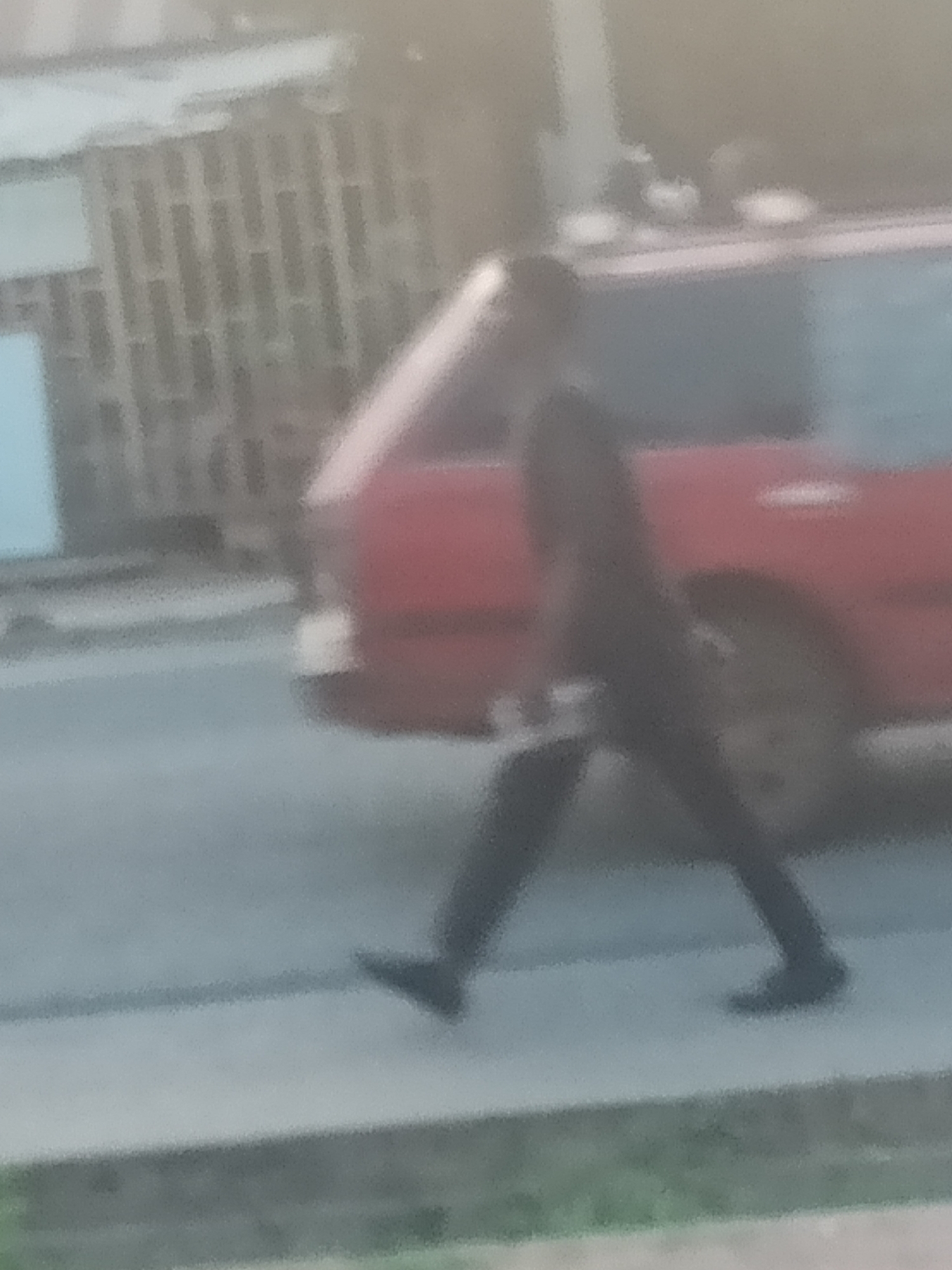 The image depicts a blurry, low-resolution scene in Jalingo, Nigeria, featuring a person walking on a paved surface. The individual, dressed in dark clothing, appears to be moving from left to right across the frame. Behind the person, a red vehicle is partially visible, suggesting a street or parking area. The background includes indistinct structures, possibly buildings or fences, with a muted color palette dominated by grays and dull reds. The overall impression is of an outdoor urban environment, though specific details are obscured by the image quality.