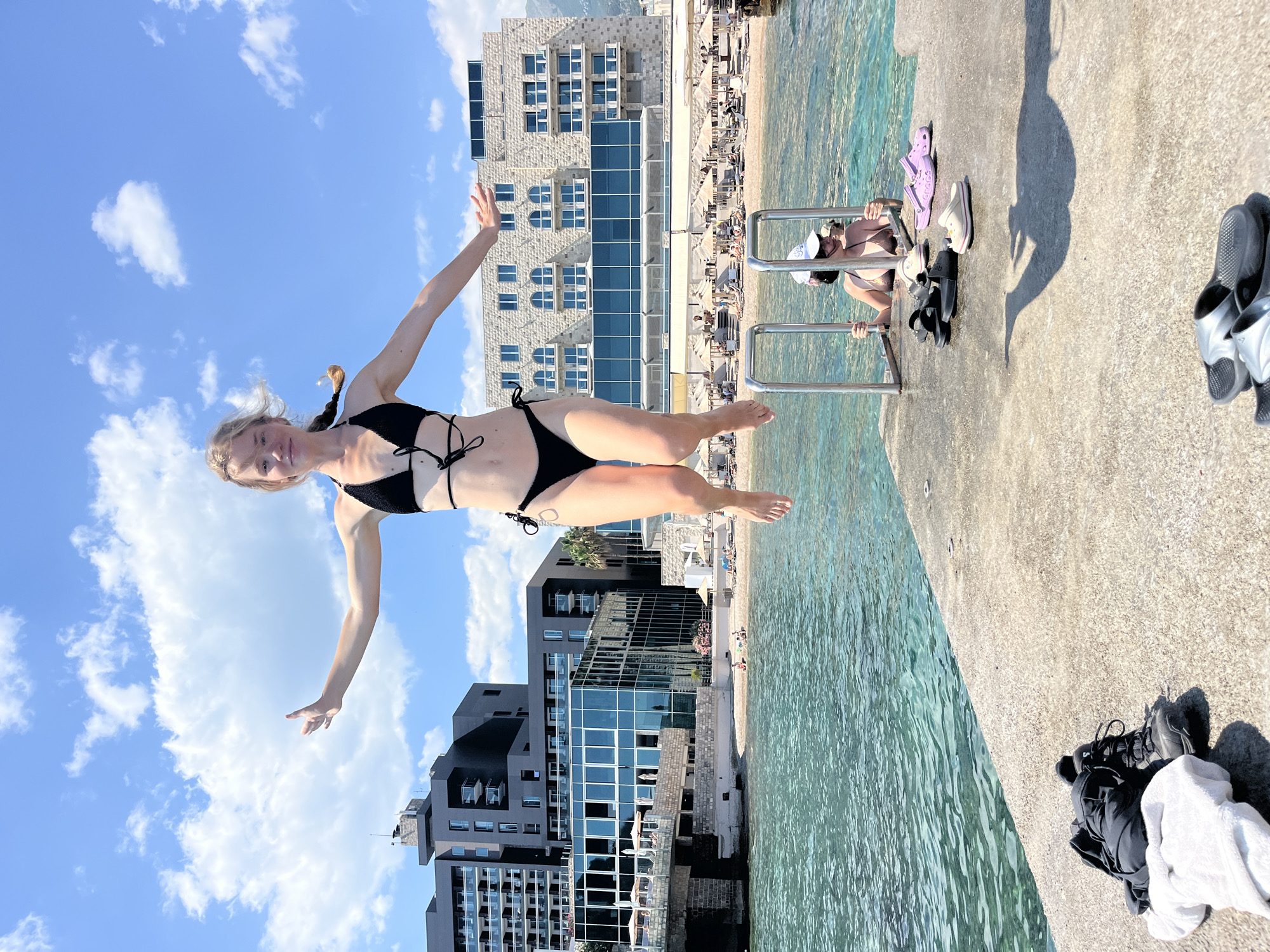 An action shot captures a young woman in mid-air, jumping off a concrete pier with the turquoise waters of the Adriatic Sea below. She is wearing a black bikini with intricate string detailing. Her arms are outstretched, and her legs are bent, showcasing a moment of exhilaration. In the background, several buildings with modern architecture and balconies line the shore. A beach with rows of umbrellas and sunbathers is visible, suggesting a popular tourist destination. Below the pier, another woman is holding onto a metal ladder, partially submerged in the water, looking up at the jumping woman. On the pier in the foreground, a black backpack and a white towel are placed near a pair of black hiking boots. Further down the pier, several pairs of colorful Crocs are scattered. The sky is a vibrant blue with scattered white clouds, indicating a clear, sunny day. The scene is likely in Budva, Montenegro, a coastal city known for its beaches and lively atmosphere.