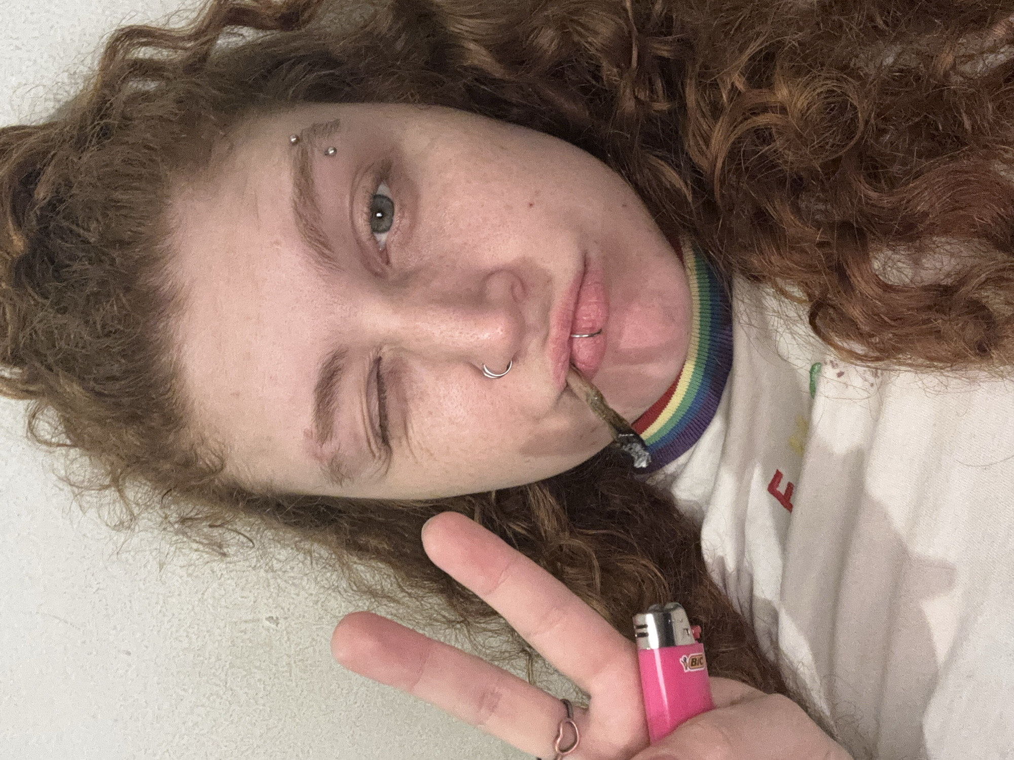 The image presents an angled, close-up portrait of a person with long, curly brown hair, light skin, green eyes, and visible freckles. The individual is shown indoors against a plain, light-colored background.
The person has multiple facial piercings, including a double piercing on their left eyebrow, a silver septum ring in their nose, and a ring on their lower lip. They are winking with their right eye (viewer's left) while their left eye remains open, conveying a playful or relaxed expression.
A hand-rolled cigarette, appearing to be lit, is held in their mouth. In the foreground, one of their hands forms a peace sign with two fingers extended. A heart-shaped ring adorns the ring finger of this hand. A pink Bic lighter, with "BIC" visible on its side, is held between the extended fingers.
The person is wearing a white collared shirt featuring a distinctive rainbow-striped pattern on the collar, suggesting an affinity for colorful fashion or LGBTQ+ pride. A partial red letter, resembling an 'F', is visible on the front of the shirt.
The image lacks explicit location or time-of-day indicators, beyond an inferred indoor setting. The overall demeanor is casual and confident.