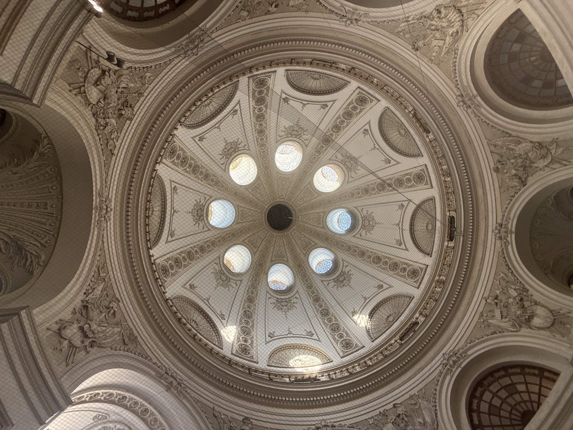 The image shows the interior of a domed ceiling, likely in a grand building. The dome features elaborate stucco work with intricate patterns and decorative elements, including floral motifs and possibly allegorical figures. Several circular windows, some with stained glass, are visible, allowing natural light to filter through. A fine mesh netting is stretched across the dome, likely for protection or to prevent debris from falling. The overall aesthetic suggests a historical and ornate architectural style, common in significant public buildings or palaces. The scene is devoid of people and focuses entirely on the architectural details of the ceiling.
This is the interior of the cupola in the central hall of the Naturhistorisches Museum (Natural History Museum) in Vienna, Austria. The museum, located on Maria-Theresien-Platz, is a monumental building with a richly decorated interior. The cupola features intricate stucco work by Josef Storck and frescoes by Hans Makart, depicting themes related to natural history and scientific exploration. The circular windows allow light to illuminate the vast space below.