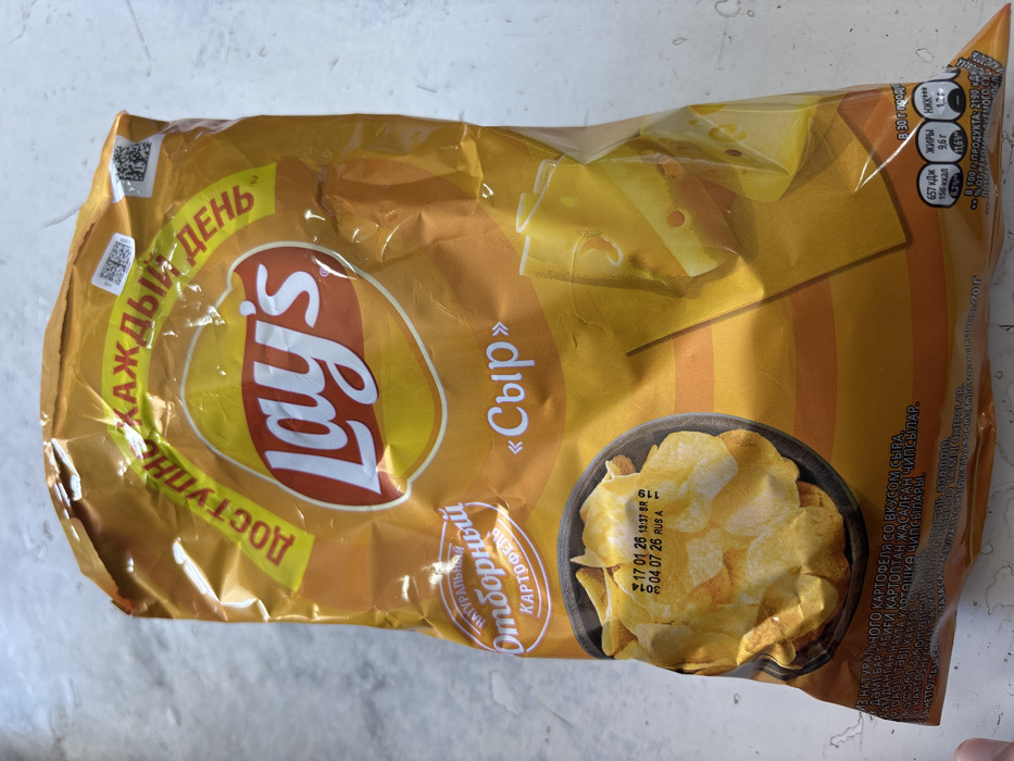 The scene depicts a single Lay's brand potato chip bag, predominantly orange and yellow, resting on a light gray, textured surface resembling a countertop. The bag is crinkled in several areas, indicating prior handling. Prominent Cyrillic text on the bag identifies the flavor as "Сыр" (Cheese) and features the slogan "ДОСТУПНЫЙ КАЖДЫЙ ДЕНЬ" (Available Every Day). Graphics include a dark bowl filled with potato chips on the lower left and several overlapping slices of cheese on the lower right. A printed date stamp on the image of the chips indicates "17 01 26 13:37 SR" (likely production date and time: January 17, 2026, 13:37) and "04 07 26 RUS A" (likely best-before date: July 4, 2026, manufactured in Russia, Batch A). Nutritional information is visible on the bottom right corner. No individuals or specific actions are discernible. The lighting is even, suggesting an indoor environment. This image is documented in the city of Lehvaz, Armenia.