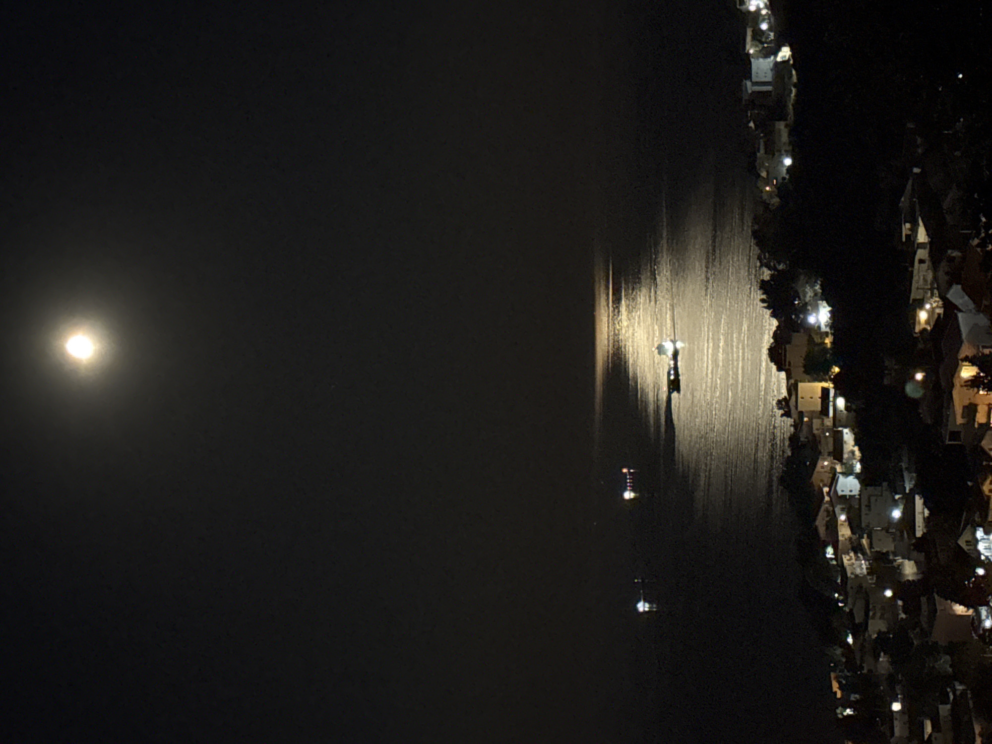 At night, a body of water reflects light, extending from the bottom to the top of the image. On the right, a coastal town is defined by closely spaced buildings and trees, illuminated by streetlights and windows. The left sky is dominated by darkness and a singular, bright moon. A tower on the water's edge reflects a light. The scene is Šušanj, Montenegro.