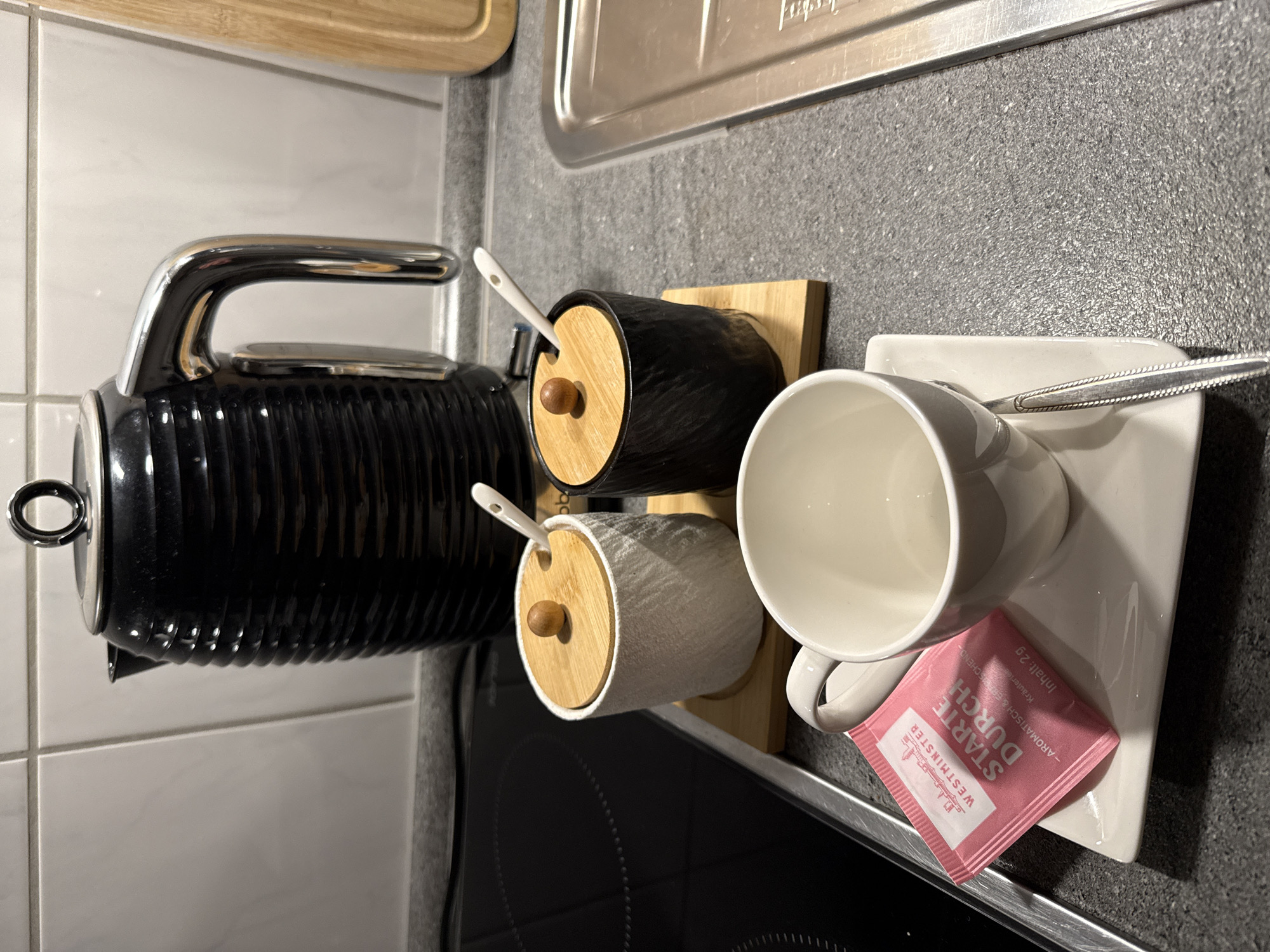 The image depicts a kitchen counter in Velbert, Germany, featuring items for preparing a hot beverage. A black ribbed electric kettle with a chrome handle is prominently positioned. To its left are two small ceramic containers with wooden lids and small spoons; one container is white and textured, the other is black and textured. A small wooden board supports these containers. In the foreground, a white teacup sits on a square saucer, with a single tea sachet, pink and labeled "WESTMINSTER STARTE DURCH", placed on the saucer. A small spoon rests on the saucer next to the teacup. The kitchen counter is grey and speckled, with a black induction stovetop visible to the left and a stainless steel sink to the right. The background shows a tiled wall. There are no people visible in the image.