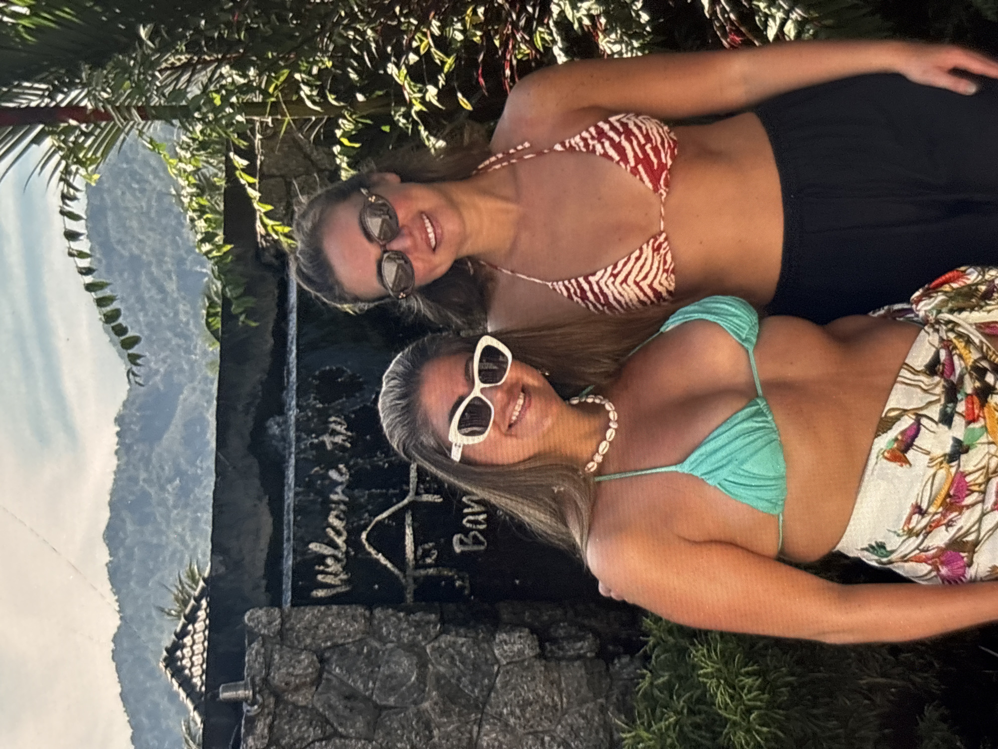 In Chalong, Thailand, two women pose in front of a stone wall with a waterfall. The woman on the left wears a turquoise bikini top and a white patterned sarong, accessorized with a shell necklace and white cat-eye sunglasses. She has long, blonde hair and smiles at the camera. The woman on the right wears a red and white striped bikini top and black pants, with oval-shaped sunglasses. She also has long, brown hair and smiles. Behind them, a sign reads "Welcome to Ban..." and a tiered mountain range is visible under a cloudy sky. Lush green foliage surrounds the scene, suggesting a tropical environment.
