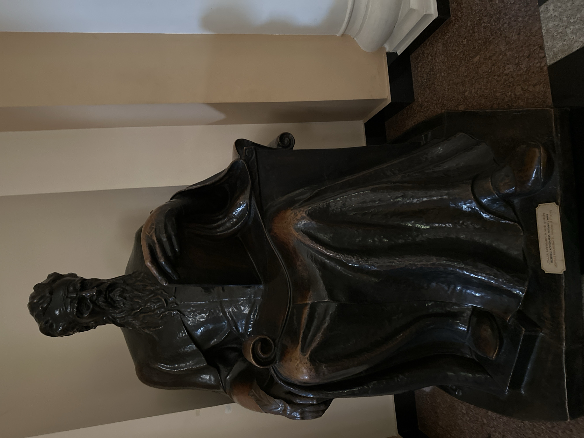 This is a full shot of a dark bronze statue of a seated man with a long beard and flowing robes, likely a historical figure. The statue is positioned in an indoor setting with light-colored walls and a textured floor. The man is holding an open scroll or book in his hands. The base of the statue has a small plaque with text that appears to be in Armenian and Latin script. The scene is well-lit, highlighting the details of the statue and its surroundings. The statue is located at the Matenadaran in Yerevan, Armenia, which is a repository of ancient manuscripts.