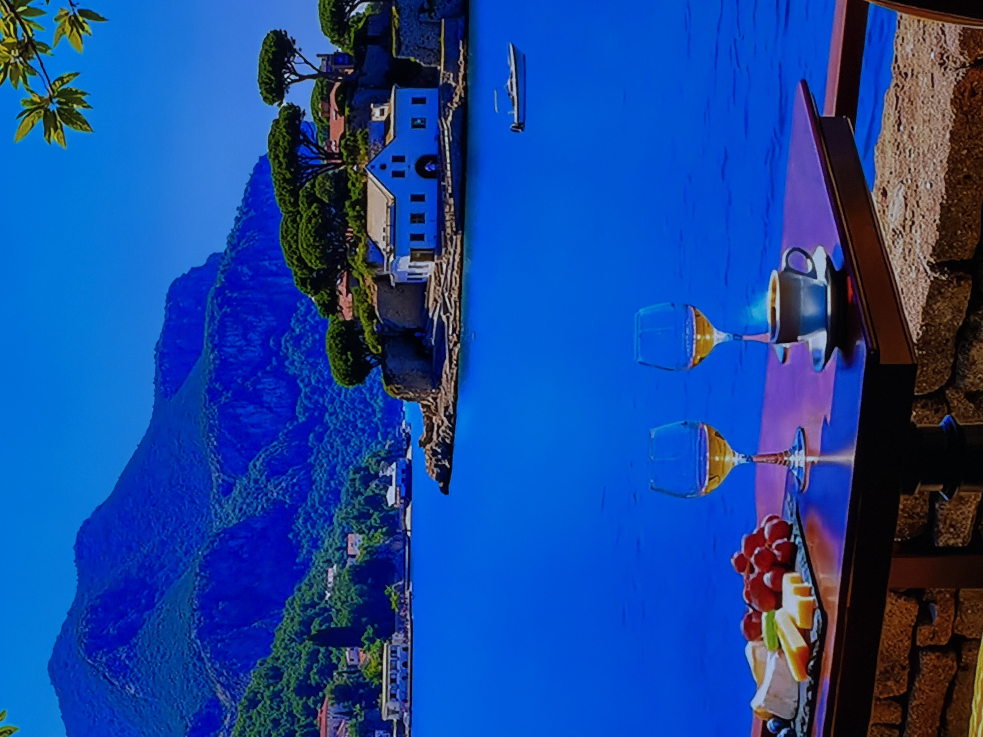 This image captures a serene, picturesque coastal scene viewed from an elevated perspective, likely a balcony or terrace. In the immediate foreground, on the right side, there is a rustic table set with a dark coffee cup on a saucer, two wine glasses (one containing a pale liquid, possibly white wine), and a rectangular platter laden with red grapes and various slices of cheese. A stone wall or railing is visible next to the table. A leafy green branch hangs into the top-left corner of the frame, adding to the natural ambiance.
The middle ground is dominated by a vast expanse of calm, deep blue sea, on which a small, white boat floats peacefully. Along the far coastline, a charming village with white and light-colored buildings, some featuring prominent windows and reddish roofs, is nestled among lush green vegetation. Distinctive pine-like trees are prominent around the buildings and along the cliffs. In the background, a majestic, rugged mountain range, appearing dark blue-green and covered with dense foliage, stretches across the left side of the scene under a clear, bright blue sky.
The time of day appears to be sunny, either morning or afternoon, with excellent visibility and calm weather. The architecture and natural landscape suggest a Mediterranean coastal location. The overall atmosphere is one of tranquility and leisure. There are no people visible in the image, nor is there any visible text.