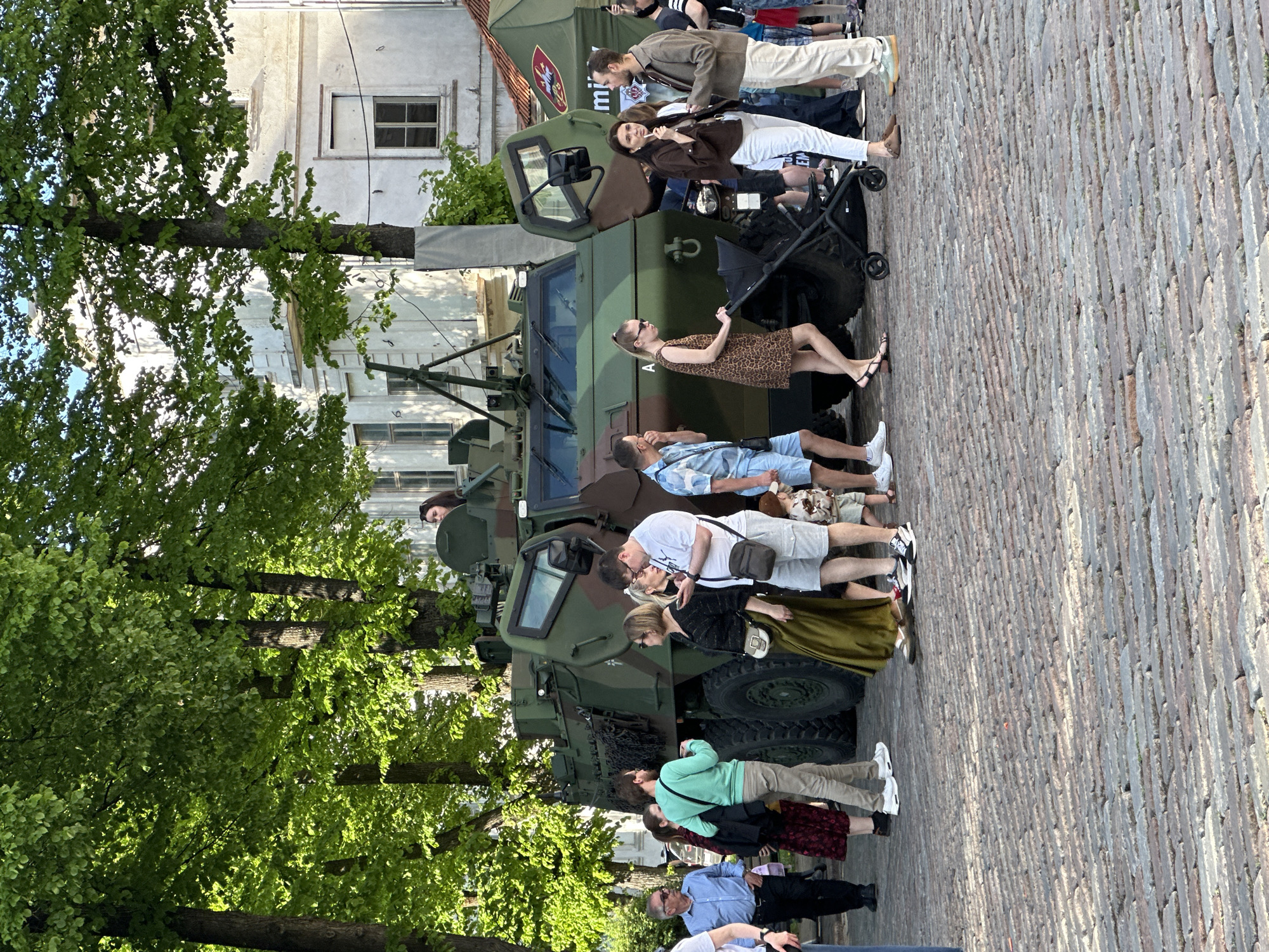 People stand near and interact with a military vehicle in what appears to be an urban environment. The vehicle is camouflage green with what may be a Latvian symbol. A group of people is seen near it, including a woman in a leopard print dress and another pushing a baby stroller with a child in it. A couple is kissing in the crowd. A building is visible in the background, along with trees on the left. The ground is paved with stone. This scene could be in Riga, Latvia, given the context of a Latvian symbol.