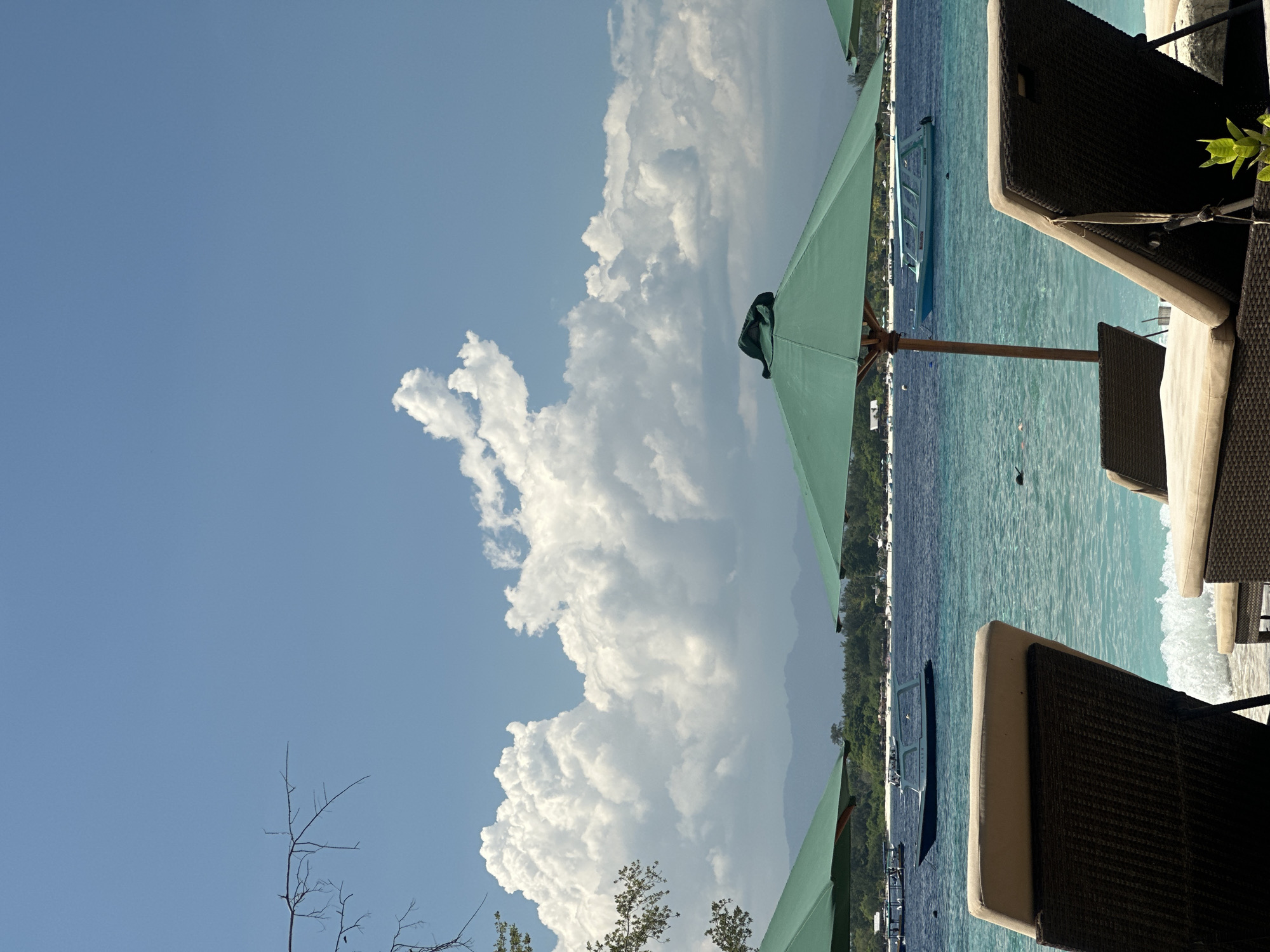 The image captures a coastal scene, likely a resort or beachfront property. The sky is predominantly blue, with a large formation of cumulus clouds dominating the view. Part of a green parasol is visible, framing the scene, and partially obscuring a view of the sea. The sea is calm, and varies from blue to turquoise. In the water, several boats are visible. The right side of the image features patio furniture, specifically chairs made of wicker material. The location is Unknown, Indonesia.