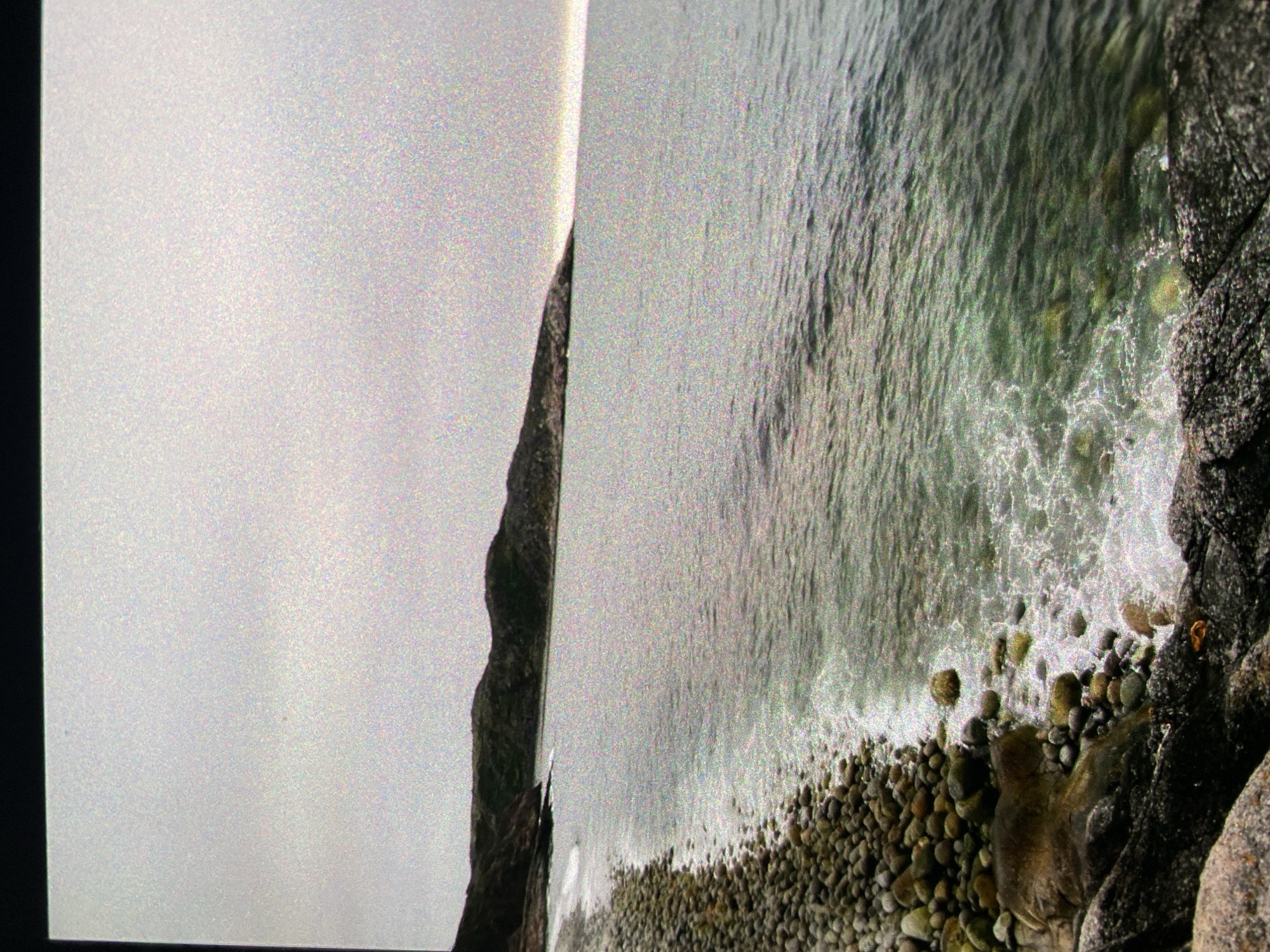 The image shows a rocky coastline with a body of water. The left side of the image is dominated by a cliff face shrouded in mist or fog. The body of water occupies the majority of the right side of the image, with waves washing onto a beach covered in pebbles and rocks. The presence of coastal rocks suggests a natural environment. Without additional information, it's impossible to confirm if this location is specifically in Saint Petersburg, Russia, as coastal features can be found in many locations worldwide. The image displays no identifiable people, actions, or interactions. The focus is on the environment and geological formations.