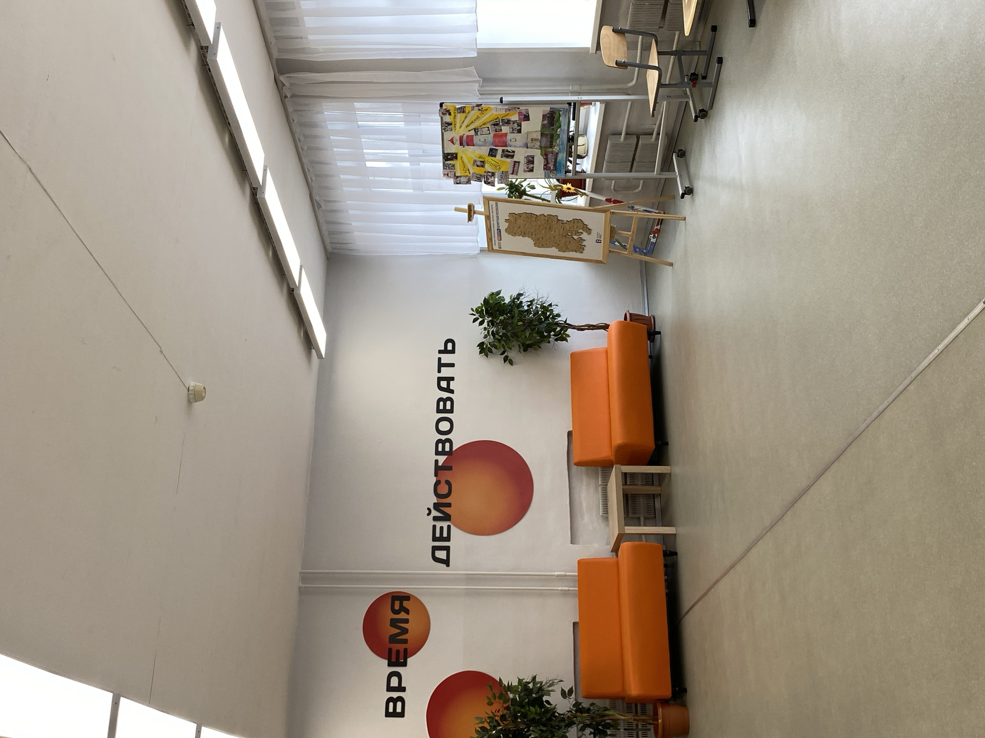 This image depicts a bright and modern indoor common area, likely within a school or educational establishment. The room features white walls and a light-colored, speckled floor, creating a clean and open atmosphere.
A prominent feature is the large Russian text on one wall, which reads "ВРЕМЯ ДЕЙСТВОВАТЬ," translating to "TIME TO ACT." This motivational phrase is accompanied by several vibrant orange and red circular graphics. The space is well-lit by a series of long, rectangular LED light panels mounted high on the walls, supplementing the natural light streaming in through large windows draped with translucent white curtains.
Furnishings include two contemporary orange sofas, each paired with small wooden side tables, suggesting an area for relaxation or informal meetings. Green potted plants are strategically placed near the seating, adding a touch of nature. Towards the back of the room, near the windows, an easel displays a wooden, relief-style map, possibly of a specific region. Beside it, another stand holds a colorful collage or poster featuring a lighthouse and various smaller images and text, one of which is visible as "МАЯК" (Lighthouse). In the background, several classroom-style desks and chairs are visible, some stacked, reinforcing the educational context.
No people are present in the image, suggesting a quiet moment in the space. The overall impression is one of a clean, organized, and inviting environment designed for students or staff, with a clear message of encouragement.