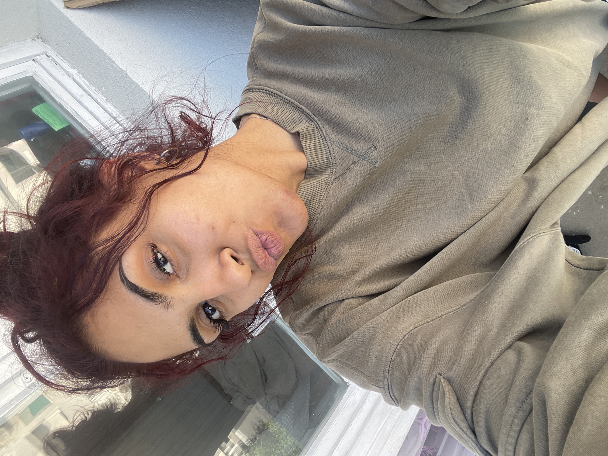 A female adult is depicted in a close-up selfie, captured at an upward angle. She has reddish-brown hair styled in a high, messy bun with loose strands framing her face and ears. Her dark eyes are open and directed at the camera, featuring defined eyebrows and long eyelashes. Her lips are puckered into a "kissing" or "duck face" expression. Visible on her right ear (viewer's left) is a small, silver-colored hoop earring, and a thin silver-colored chain necklace is present around her neck. She wears a plain, muted olive-green or taupe long-sleeved crew-neck sweatshirt with a ribbed collar, cuffs, and hem. A distinctive X-shaped stitch is visible below the front of the collar.
The background features a white-framed window located behind her and to the upper right. Reflections in the window glass show indistinct outdoor elements, including what appear to be distant buildings and greenery, along with partial reflections of an interior space (including a blue and green object). A light-colored, possibly off-white or light grey, textured wall is visible adjacent to the window frame. The scene is illuminated by natural light, likely from the window, casting subtle shadows on her neck and the wall. The subject appears to be indoors in the city of Unknown, Unknown.