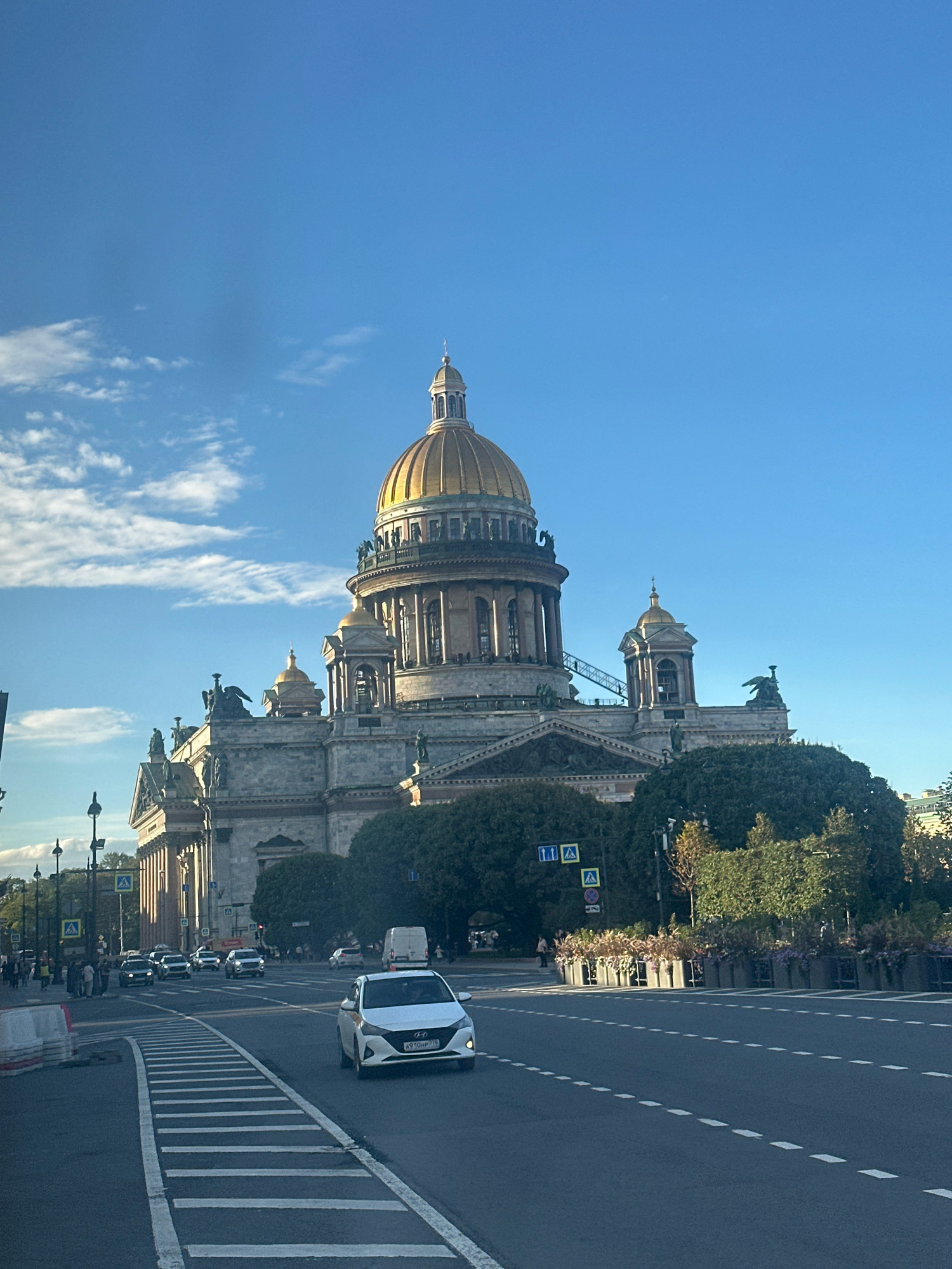 This is a daytime, eye-level, outdoor photograph of St. Isaac's Cathedral in Saint Petersburg, Russia. The massive, neoclassical cathedral, with its prominent golden dome and intricate stonework, dominates the background. In the foreground, a white taxi is driving on a multi-lane road, its headlights on. The road is marked with white lines. Pedestrians are visible on the sidewalk on the left side of the image, and a traffic light is visible. Trees and flower planters line the side of the road. The sky is blue with scattered white clouds. The overall impression is of a bright, sunny day in a large European city.