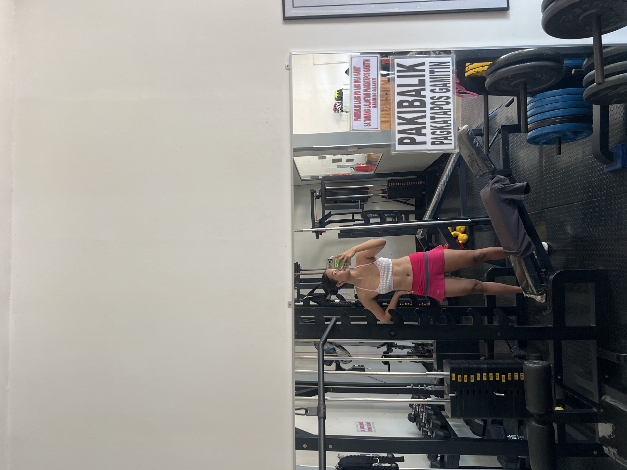 In the city of Cuyo, Philippines, a young woman in athletic wear is captured in a mirror selfie at a gym. She is wearing a white and pink checkered sports bra and a pink athletic skirt. Her hair is pulled back in a bun. The gym is equipped with various weightlifting machines, including a stack of weight plates and barbells visible in the foreground and background. A sign with Filipino text, "PAKIBALIK PAGKATAPOS GAMITIN," which translates to "Please return after use," is prominently displayed on a wall. The lighting suggests it is daytime, and the overall environment appears to be a functional, well-maintained fitness facility.