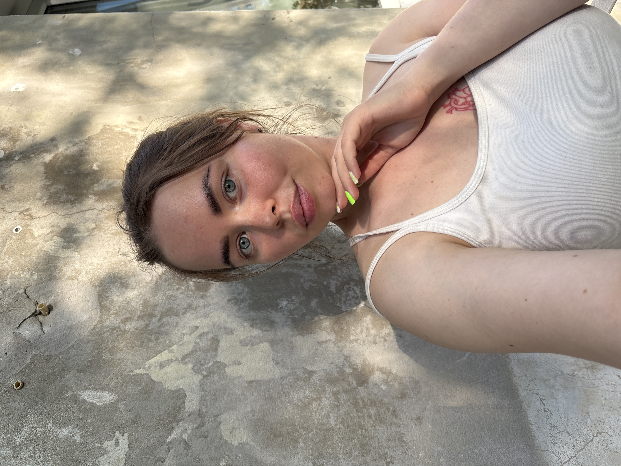 A young woman with light skin, brown hair, and blue eyes is lying on a concrete surface. She is wearing a white tank top. A small red tattoo is visible on her chest. Her right hand is near her neck, and her fingernails are painted neon green. The background is a gray concrete surface with some debris. The location is not discernible from the image, therefore the inclusion of Izhevsk, Russia is not possible.
