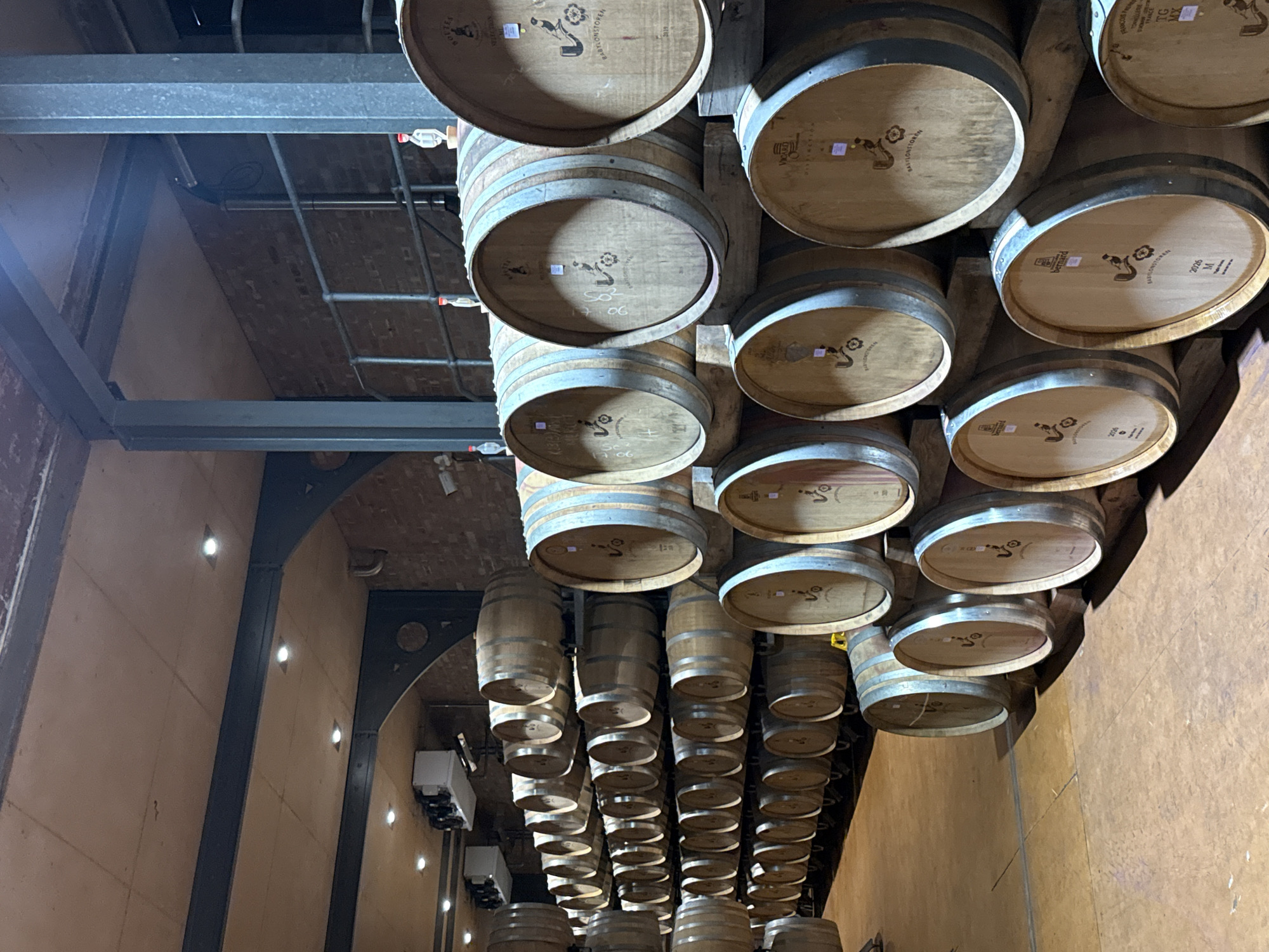 This image captures the interior of a wine cellar, showcasing numerous wooden wine barrels meticulously stacked and stored. The perspective is tilted, looking upwards towards the ceiling where many barrels appear to be suspended or supported by a robust metal beam structure. The barrels are arranged in multiple rows and layers, extending deep into the facility.
The scene is dominated by the warm, earthy tones of the wooden barrels and the light brown walls, contrasted by the grey metal beams and piping. The walls feature sections of exposed brickwork and smoother, lighter-colored surfaces with small, embedded light fixtures illuminating the space. On the left side of the image, what appear to be refrigeration or cooling units are mounted on a wall. Many of the barrels have small, clear plastic containers or airlocks attached to their tops, indicating active fermentation or the careful aging process of wine.
Notable details include the visible text and logos on the barrel ends. A prominent logo depicting a stylized figure is accompanied by the word "BABYLONSTOREN," clearly identifying the location as Babylonstoren, a wine estate in Drakenstein Nu, South Africa. Other legible text includes "SO," "17 06," "Bernard," "2023 M," "VOO," "TG MX," "Francois," and "DISTINCTION," likely referring to barrel makers, vintage years, batch numbers, or specific wine characteristics. The setting is an indoor, industrial-yet-traditional space, designed for the quiet maturation of wine. No people are visible, and the scene evokes a sense of organized production and the extensive process of winemaking.