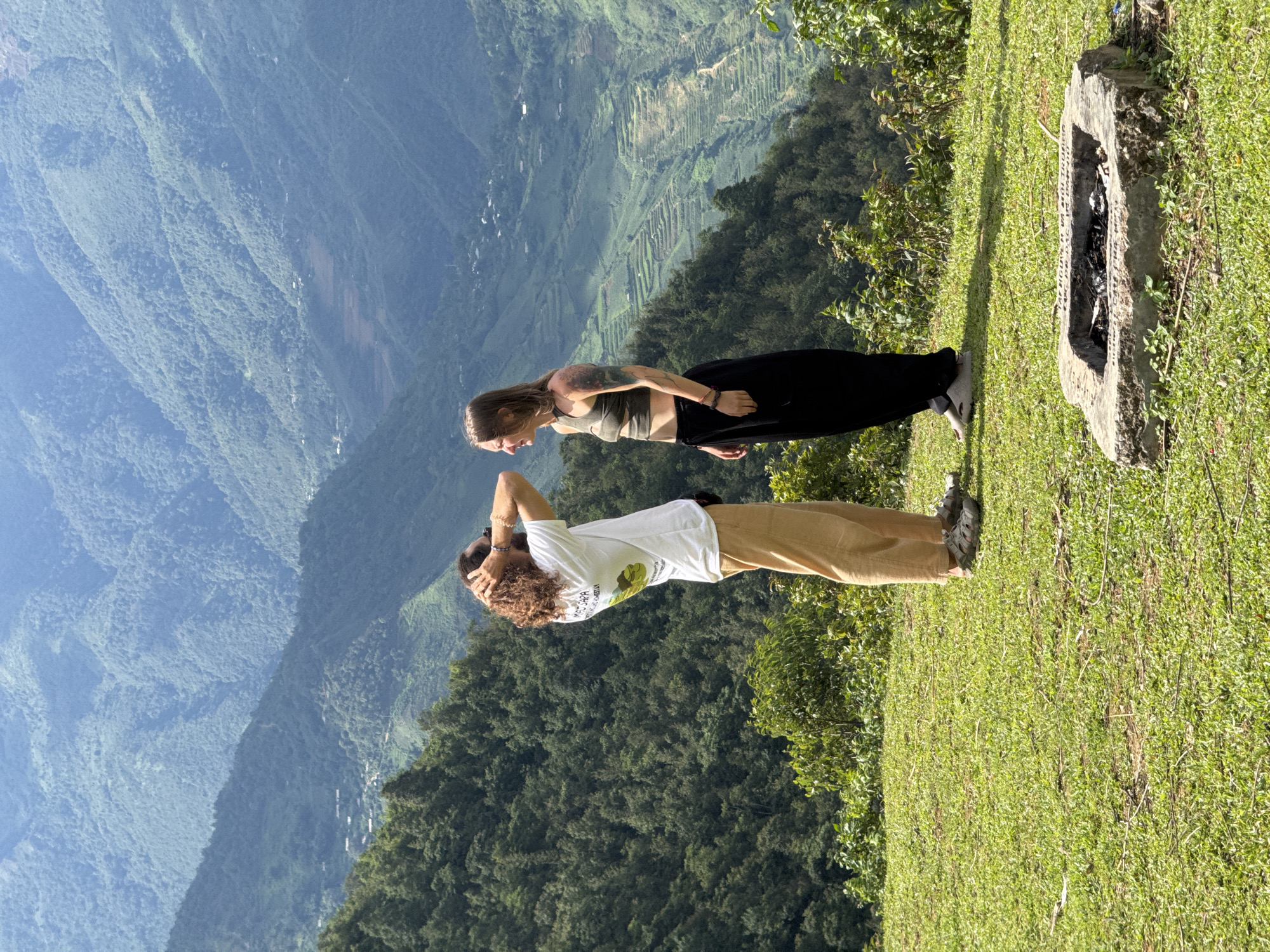 The image depicts two individuals standing on a grassy hill overlooking a vast mountainous landscape in Tu Le, Vietnam, during daylight hours.
On the left, a male with long, curly brown hair and a beard wears a white short-sleeved t-shirt with "MAI SAPA" and a graphic visible on the back, light brown pants, and grey sandals. His hands are placed behind his head, and he is looking slightly to his right, towards the other person.
On the right, a female with straight, shoulder-length light brown hair and tattoos on her left arm wears a green patterned crop top, black loose-fitting pants, and light-colored athletic shoes or sandals. She stands facing the male, with her head tilted, looking at him with a slight smile.
The ground in the foreground is covered in green grass with minimal scattered debris. In the lower right corner, a square, rough-hewn concrete fire pit containing ashes and refuse is embedded in the grass. Inscriptions are present on its visible sides.
The midground behind the individuals consists of densely forested hills. The background features multiple layers of mountains covered in green vegetation, receding into a light blue atmospheric haze. Numerous rice terraces are distinctly visible on the slopes of the closer mountains, and small clusters of buildings indicative of villages are scattered across the valleys and hillsides. The scene is brightly lit by natural sunlight.