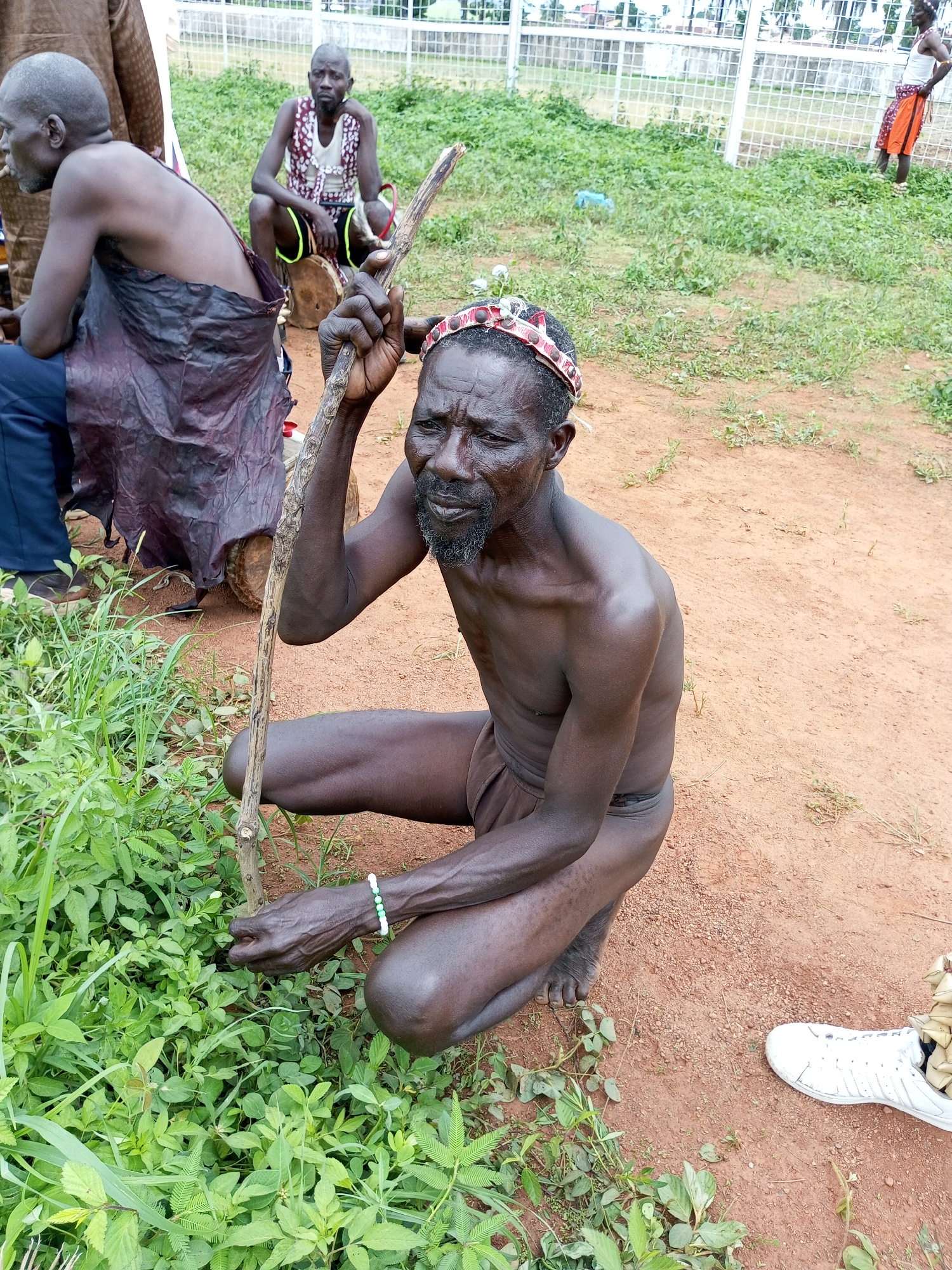 The image captures a scene outdoors, likely in Takau, Nigeria, given the prompt context. The foreground features a dark-skinned man crouching on a reddish-brown dirt surface with patches of green vegetation. He is holding a wooden stick and wearing a decorative headband and a bracelet. In the background are other individuals, including one seated on a drum-like object and another standing near a white fence. A pair of white sneakers is partially visible on the lower right side of the image.