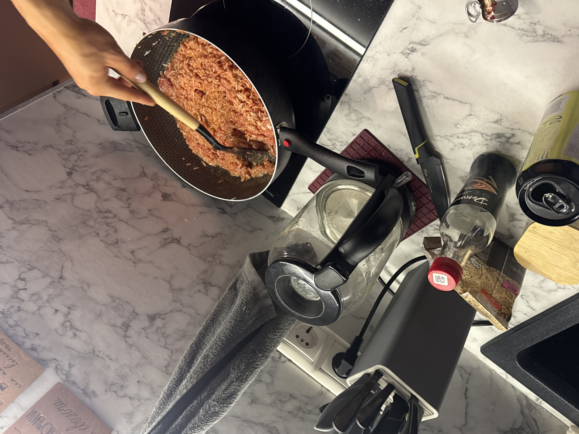 The scene is a kitchen counter. A hand holds a cooking utensil in a frying pan filled with a mixture of minced meat and rice. The frying pan is on a cooktop. Beside the cooktop, there are a knife, a bottle of spices, a beverage can, a wooden cutting board, a small bottle with a red cap, a container with kitchen utensils, a kettle, a gray dishcloth, and decorative kitchenware items. The location is a kitchen, possibly in a residential setting in Khimki, Russia.