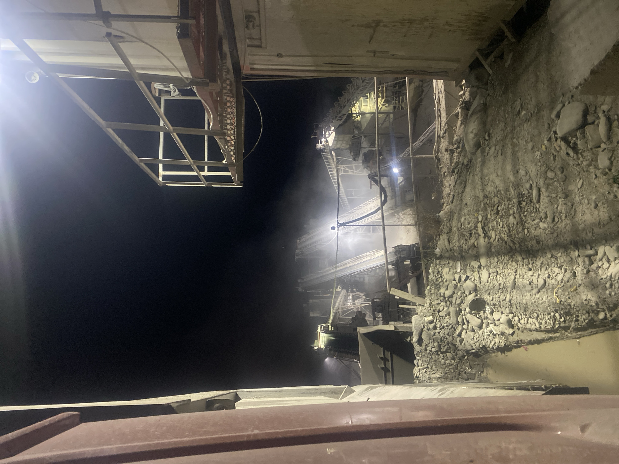 The image depicts a nighttime scene at a quarry or industrial facility, possibly a stone crushing plant, in Kharovsha, Azerbaijan. The foreground is dominated by a large, reddish-brown metallic structure on the left, appearing to be part of a vehicle or industrial equipment. Adjacent to this is a lighter-colored wall or structure, also metallic. A walkway with a metal railing and a bright light overhead is visible on the right, extending into the scene. Beyond this, the industrial complex is illuminated by artificial lights, revealing conveyor belts, machinery, and piles of rocks and debris. Dust or steam is visible in the air, suggesting ongoing operations. The ground is uneven and covered with rocks and dirt. The sky is dark, indicating nighttime. There are no visible people in the frame.