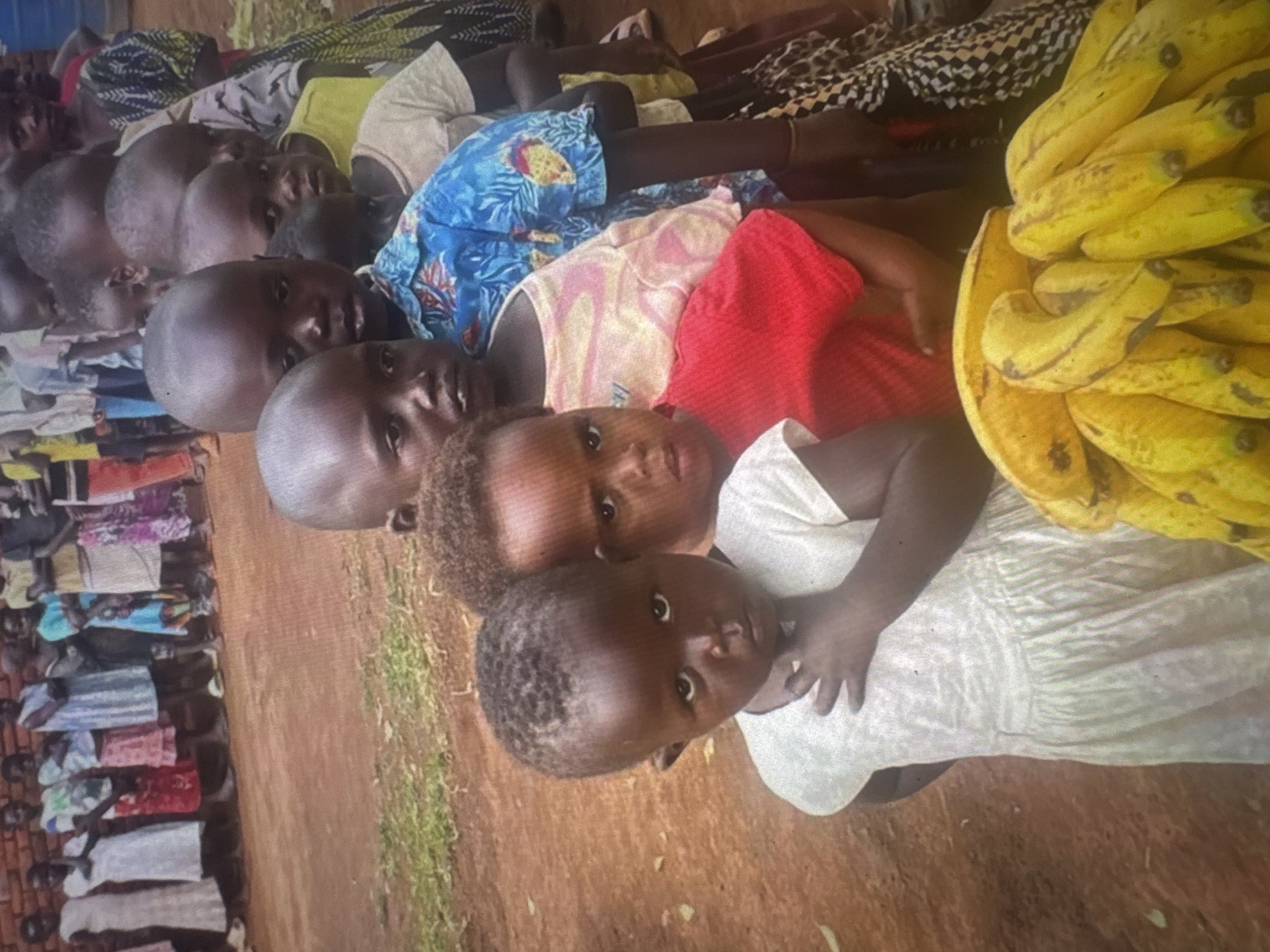 In the foreground of the image, a group of young children stand in a line, facing forward. The child closest to the camera, on the left, is wearing a white dress and looking directly at the viewer with a somber expression. To their right, another child in a red t-shirt is partially visible, their face obscured. Behind them, a line of children extends into the background, some wearing colorful clothing and others in more subdued tones. In the very front, a bunch of ripe bananas are arranged. The background consists of a reddish-brown dirt ground and a brick wall. The overall scene appears to be an outdoor gathering of children, possibly at a school or community event. The lighting suggests it is daytime. The specific location in Abuja, Nigeria, is not discernable from the visual information provided.