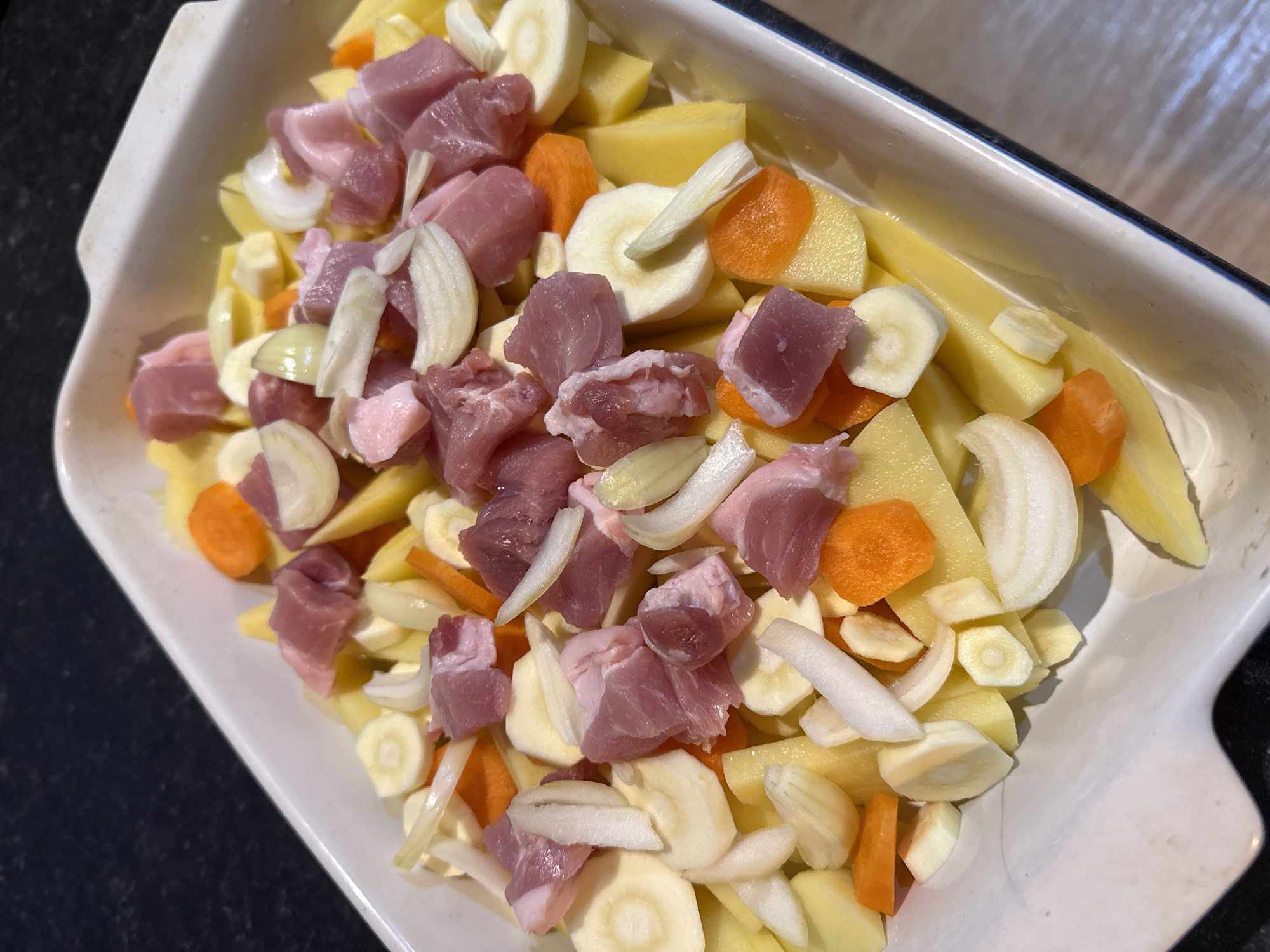 The image shows a white ceramic baking dish filled with a raw mixture of diced meat, potatoes, carrots, onions, and parsnips. The vegetables are cut into various shapes: the potatoes into large chunks, the carrots into thin slices, the onions into slivers, and the parsnips into thick rounds. The meat is cut into roughly equal-sized cubes. The dish appears to be sitting on a counter or table. The scene is not located in Great Yarmouth, United Kingdom.